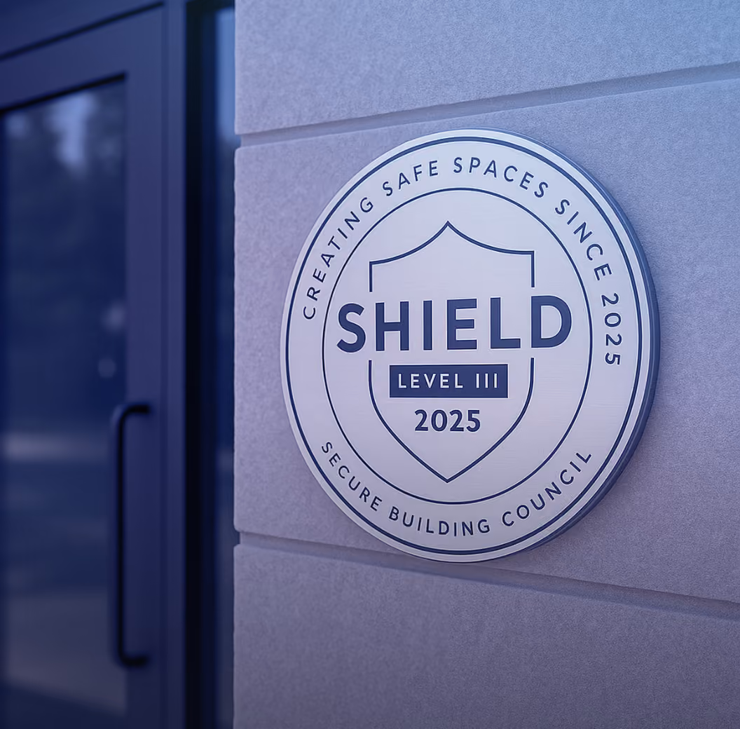 Round plaque mounted on a gray wall next to a glass door, displaying a shield emblem with the text 'SHIELD LEVEL III 2025' and the phrases 'CREATING SAFE SPACES SINCE 2025' and 'SECURE BUILDING COUNCIL' around the edge.