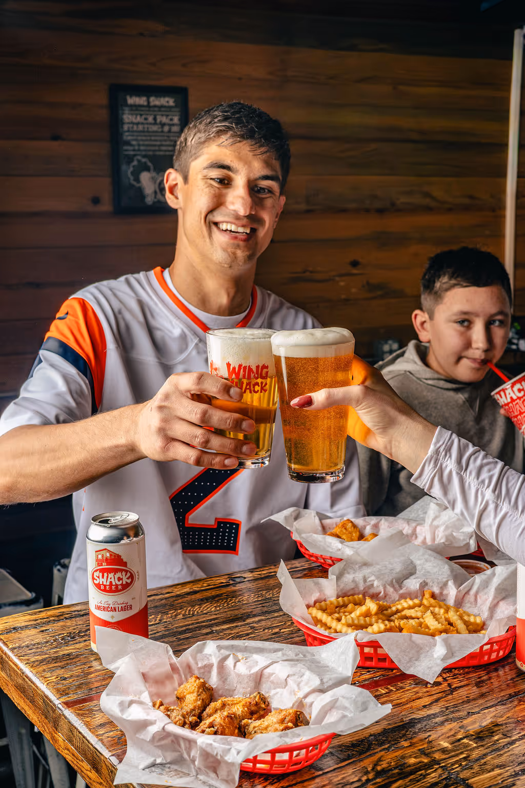 Two people holding beers from the Colorado restaurant Wing Shack with a table full of fries and wings around them.