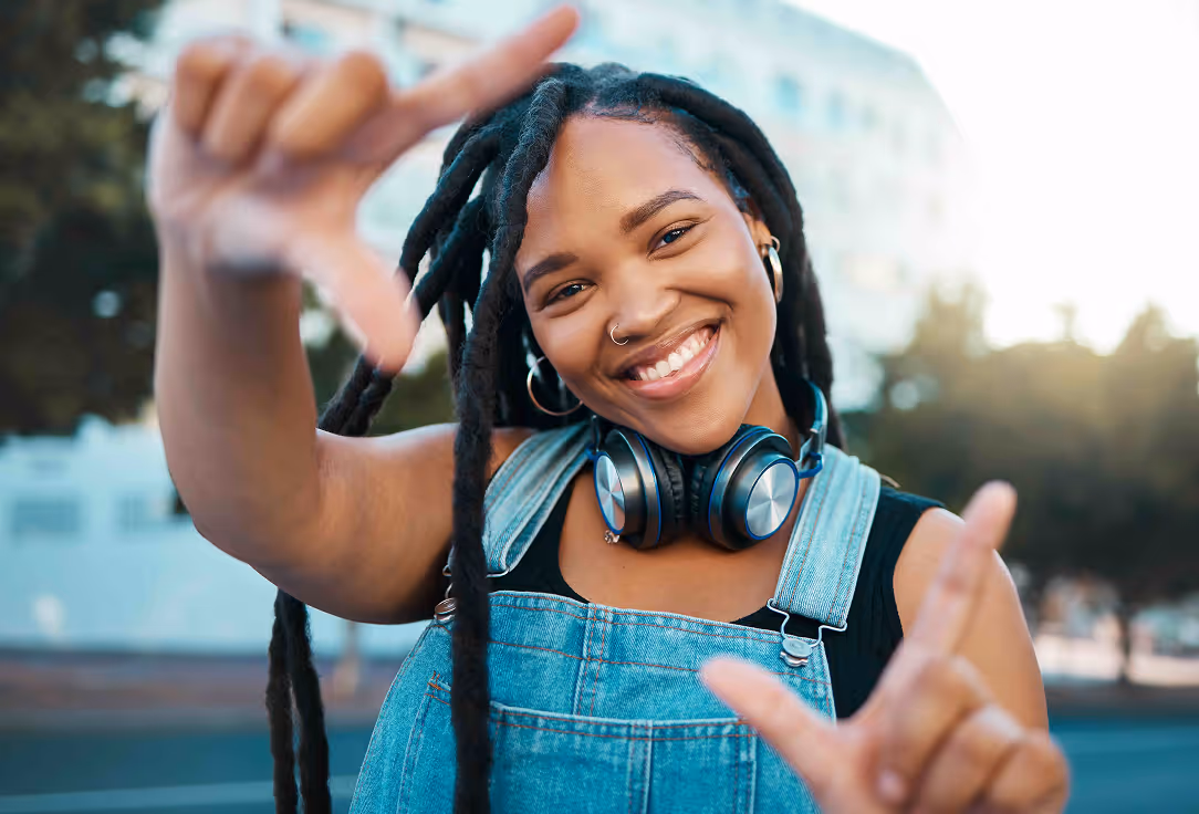 A girl wearing denim overalls and headphones makes a picture frame shape with her hands outdoors.