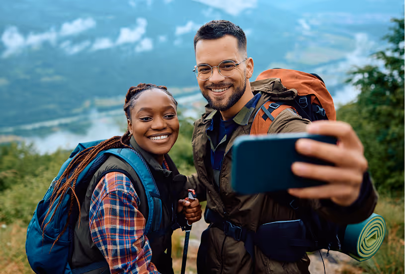 Two hikers standing on a mountain summit, taking a selfie together with scenic views in the background.