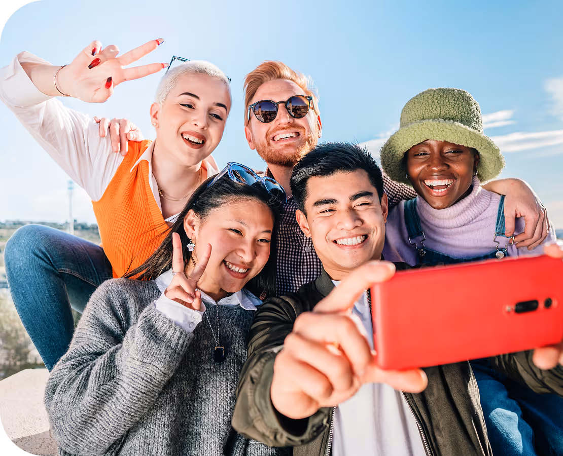 A group of friends posing together outdoors, making peace signs and taking a selfie with a red smartphone on a sunny day.
