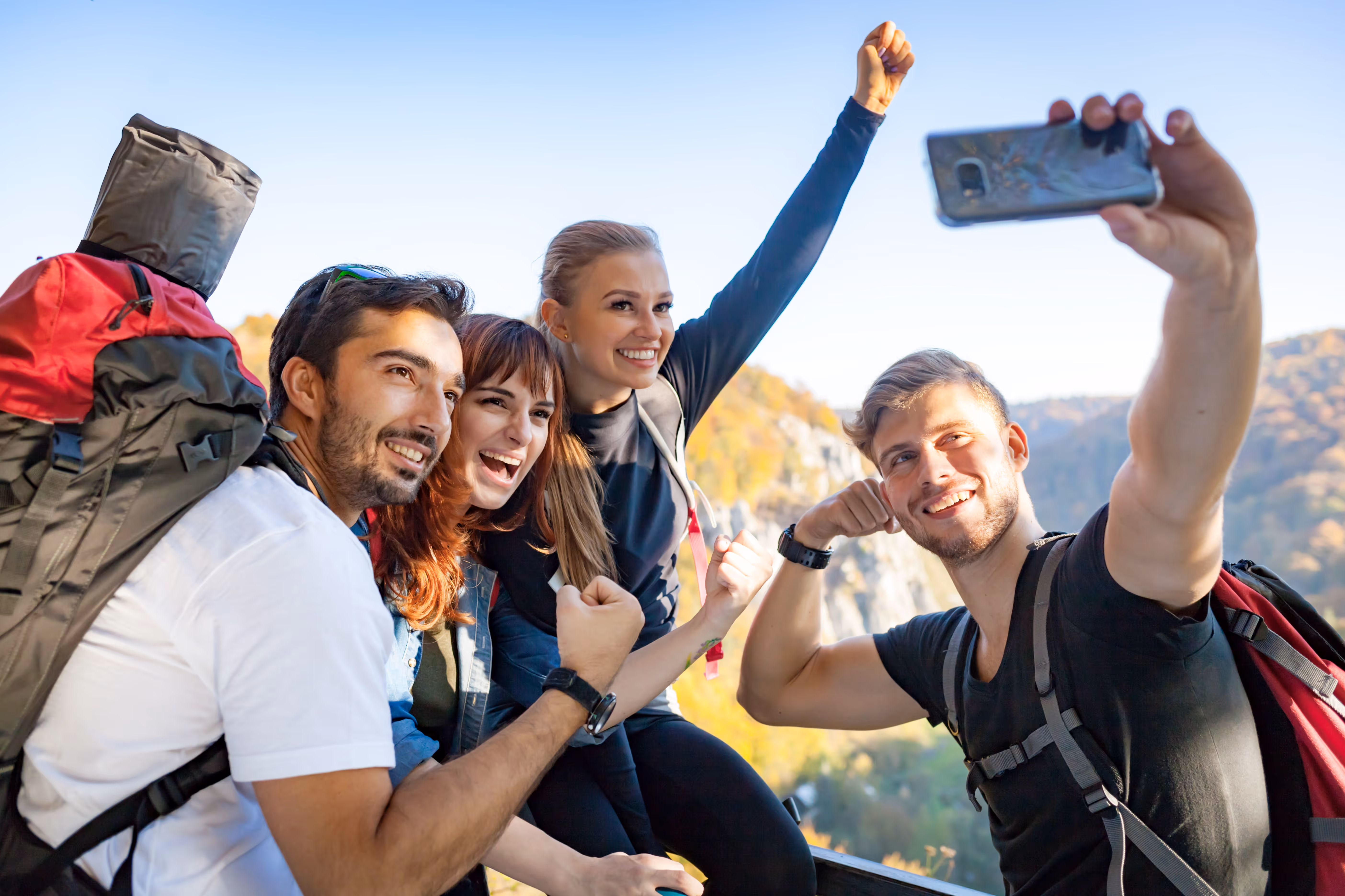 A group of hikers with backpacks posing for a selfie on a mountain trail, celebrating and flexing their arms with scenic views in the background.
