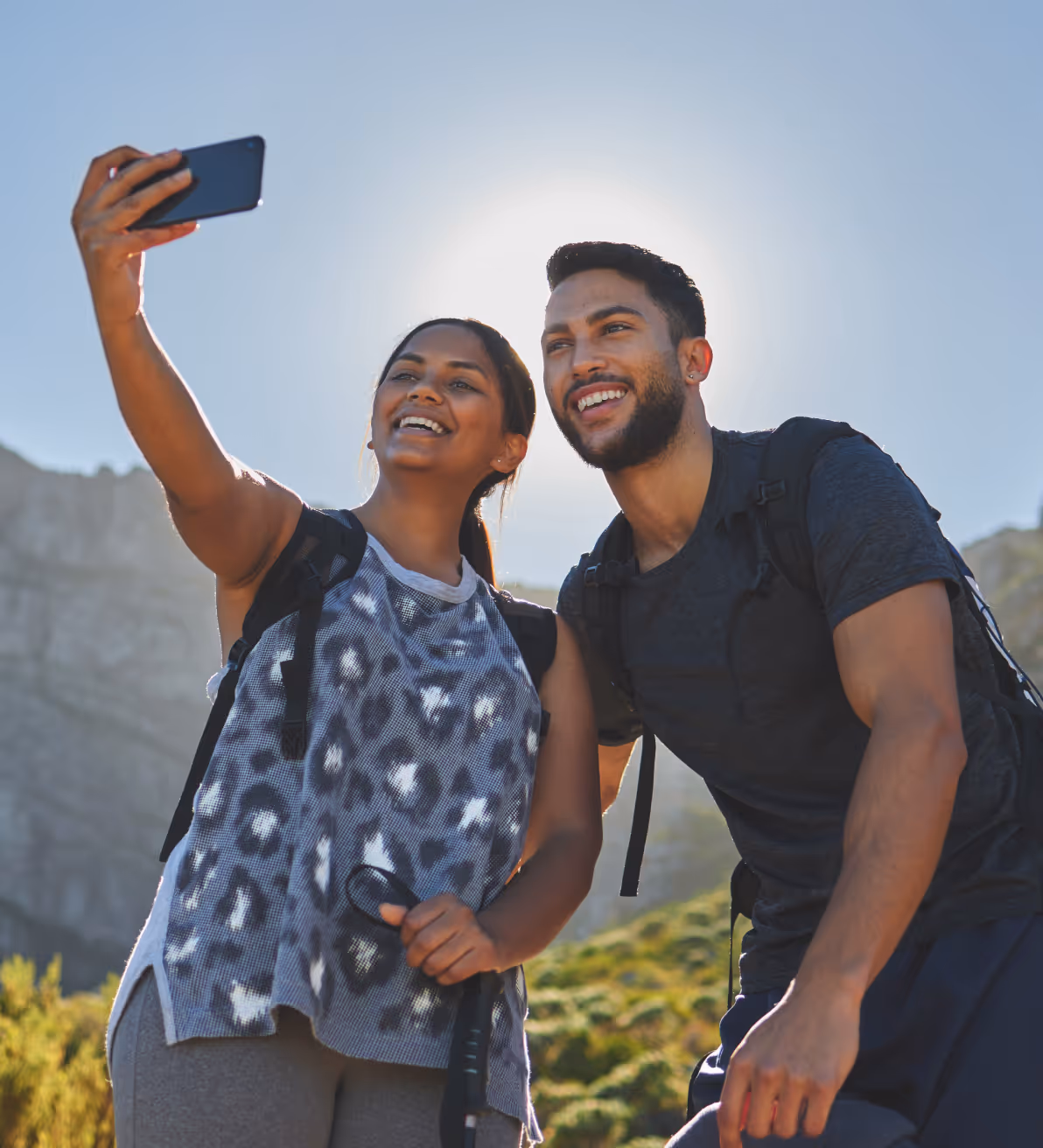 Two hikers wearing backpacks take a selfie together outdoors with mountains and greenery in the background.