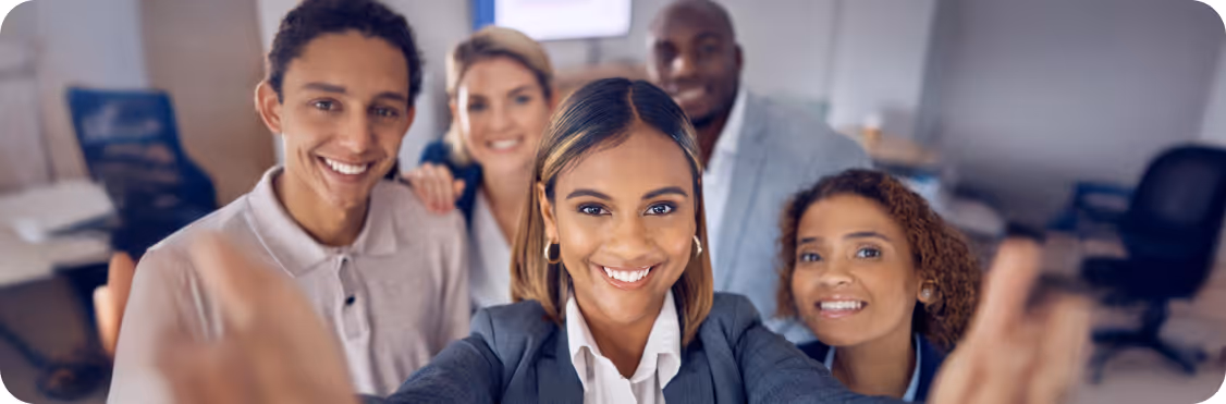A group of colleagues gathered together in an office, taking a selfie and smiling towards the camera.
