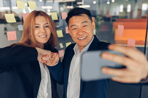 Two colleagues in business attire giving a fist bump and taking a selfie in front of a glass wall covered with colorful sticky notes in an office setting.