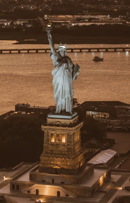 Statue of Liberty at dusk with city lights and water in the background.