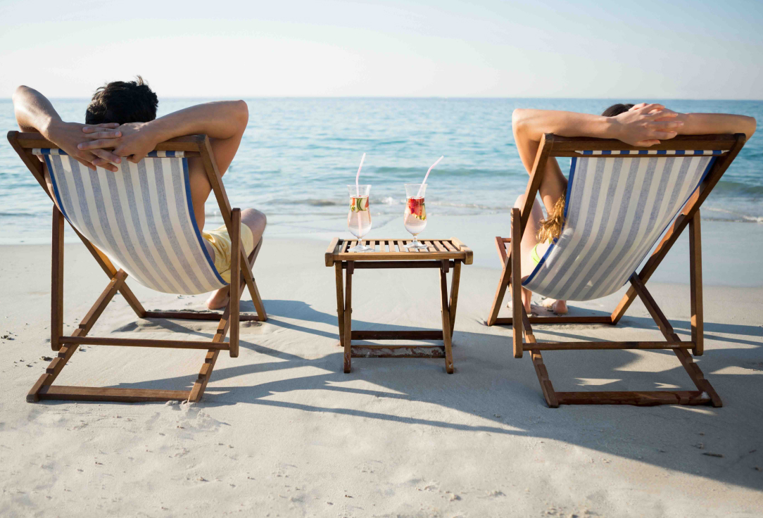 Two people relaxing on colorful beach chairs by the ocean, with a straw bag and sun hat, enjoying a sunny day with mountains and blue sky in the background.