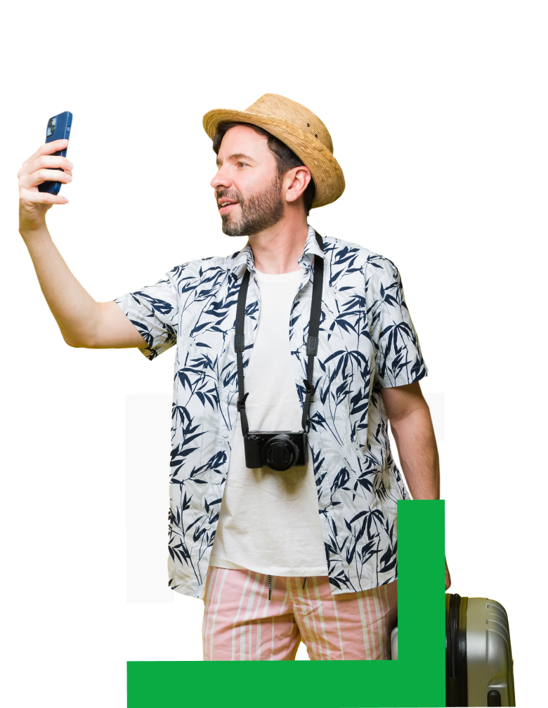 A person wearing a straw hat, tropical shirt, and shorts takes a selfie while holding a suitcase and a camera around their neck,