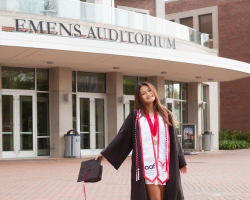 Janie posing casually in front of auditorium on Ball State's campus in her graduation gown.
