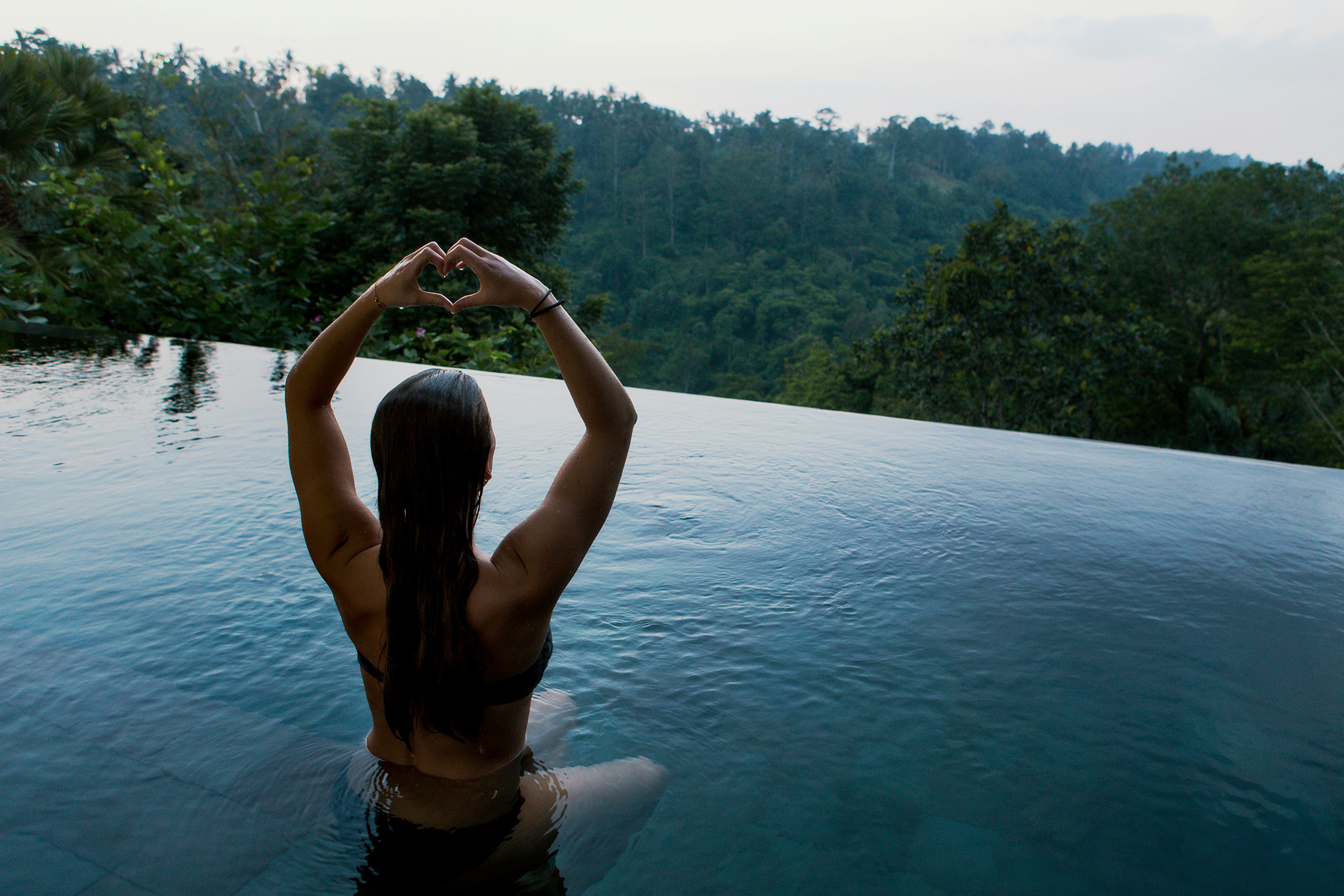 Woman holding up hands in a heart shape while sitting in an infinity pool.