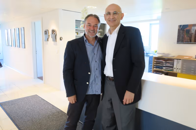 Two men standing together in a bright modern office interior, both smiling at the camera.
