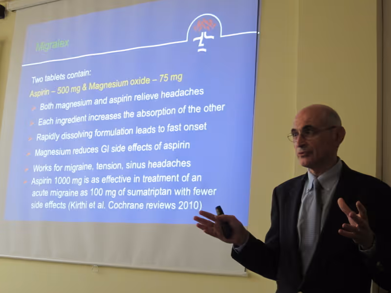 Dr. Mauskop in a suit giving a lecture in front of a projected slide presentation about migraine treatment.