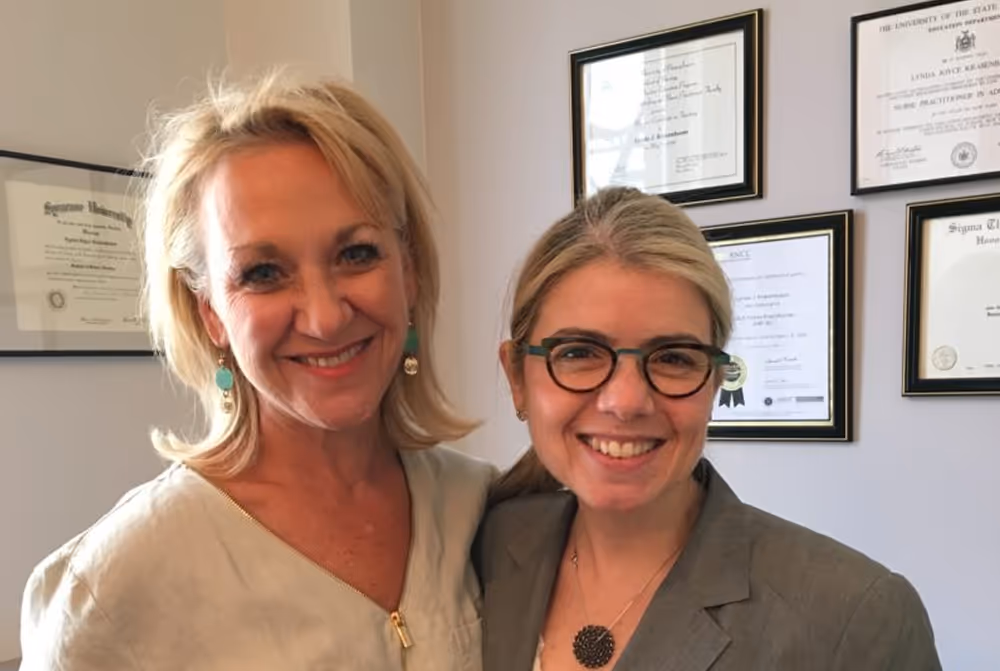 Two women smiling and standing together indoors, one wearing a white blouse and the other wearing glasses with framed diplomas on the wall behind them.