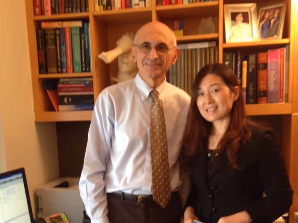 Man in glasses and tie standing next to a woman in a black jacket, both smiling indoors.