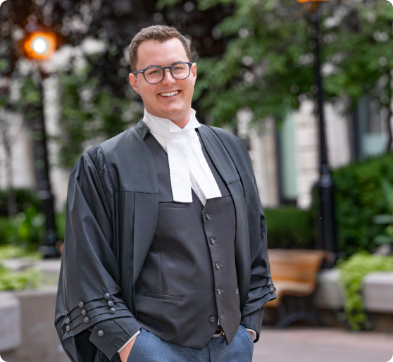 Smiling young man wearing glasses, a black barrister robe, and white neckbands standing outdoors with hands in pockets.
