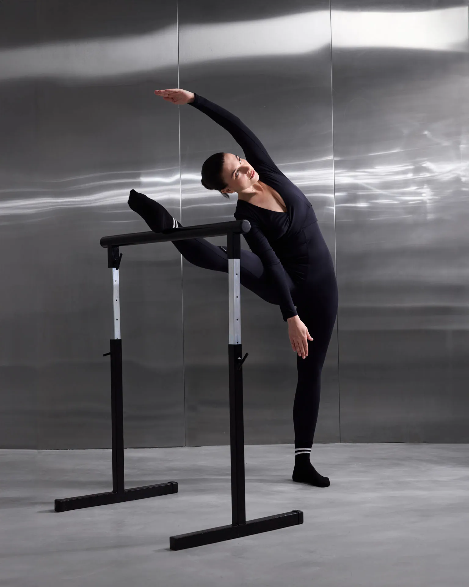 Woman in black leotard stretching leg on ballet barre in modern dance studio with metallic walls.