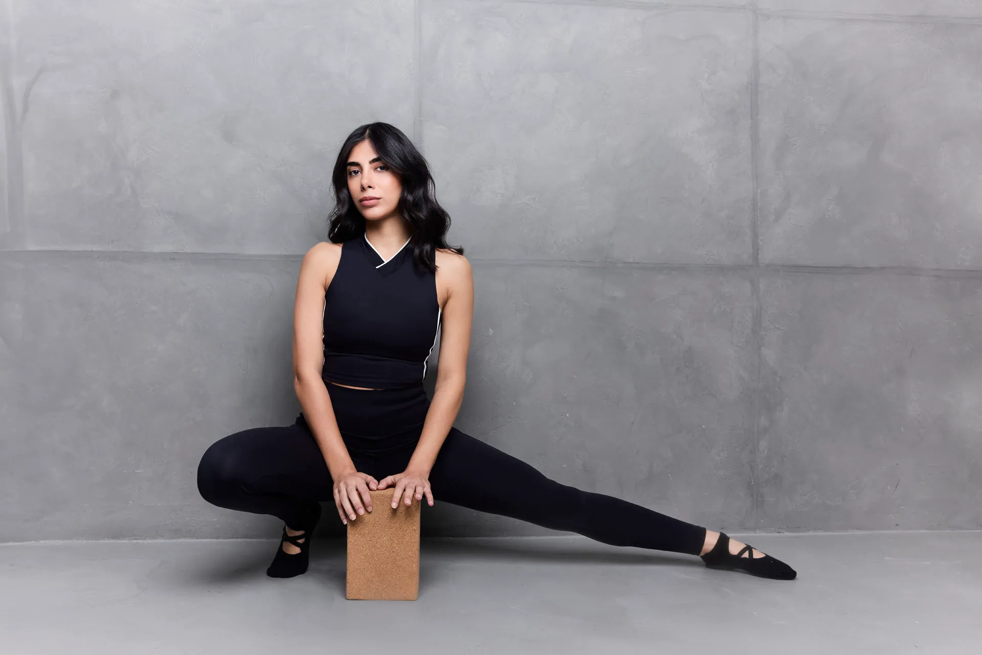 Woman in black workout clothes stretching one leg sideways while holding a cork yoga block.