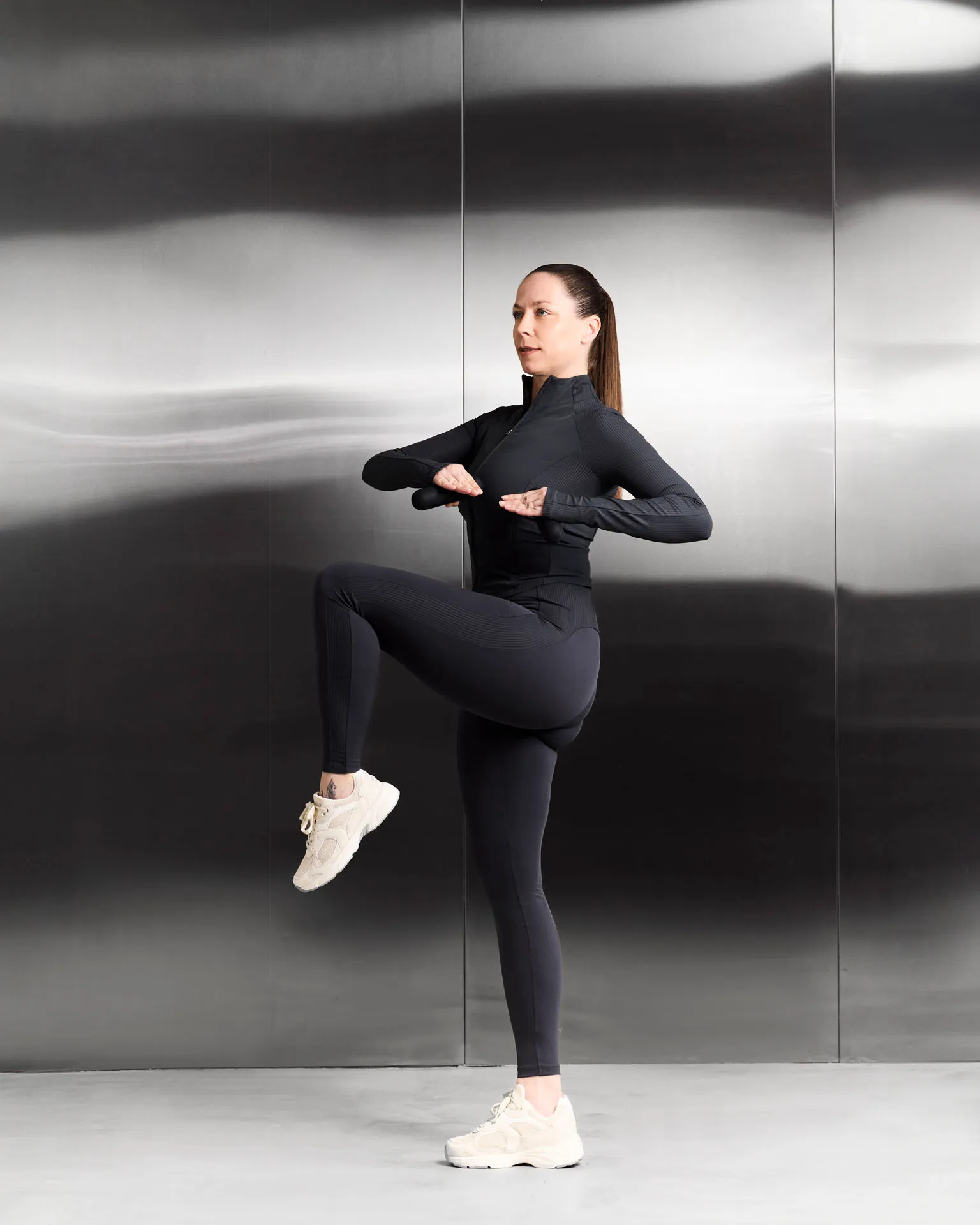 Woman in black activewear lifting one knee and holding hands in front of chest against metallic wall.