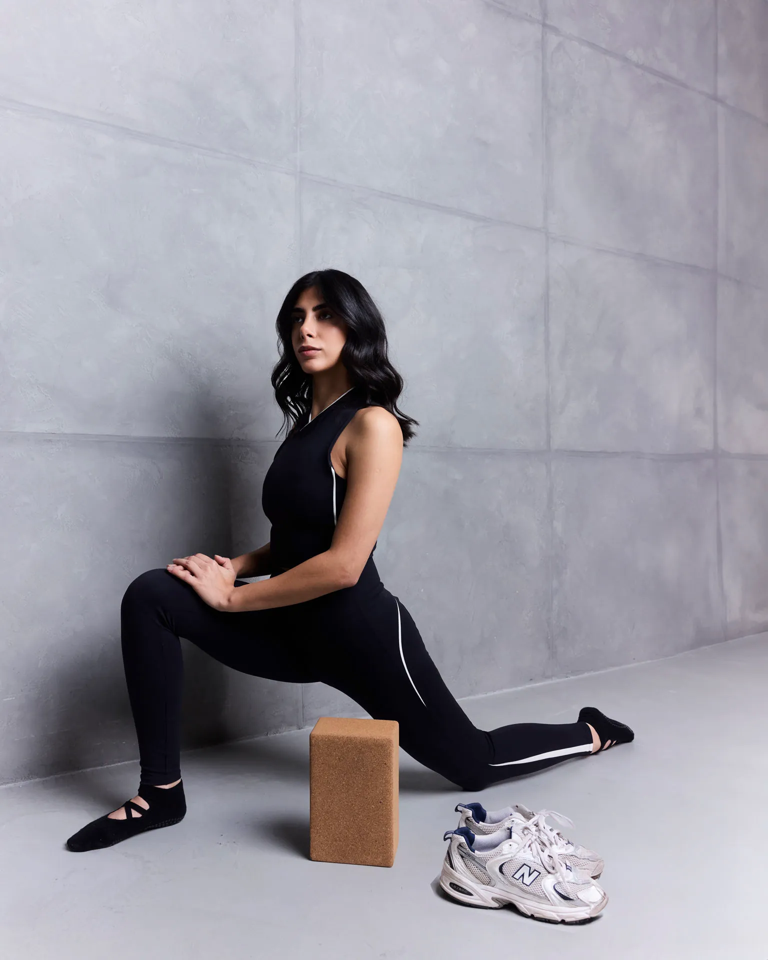 Woman in black workout clothes stretching with one knee bent and hands on knee near a cork yoga block.