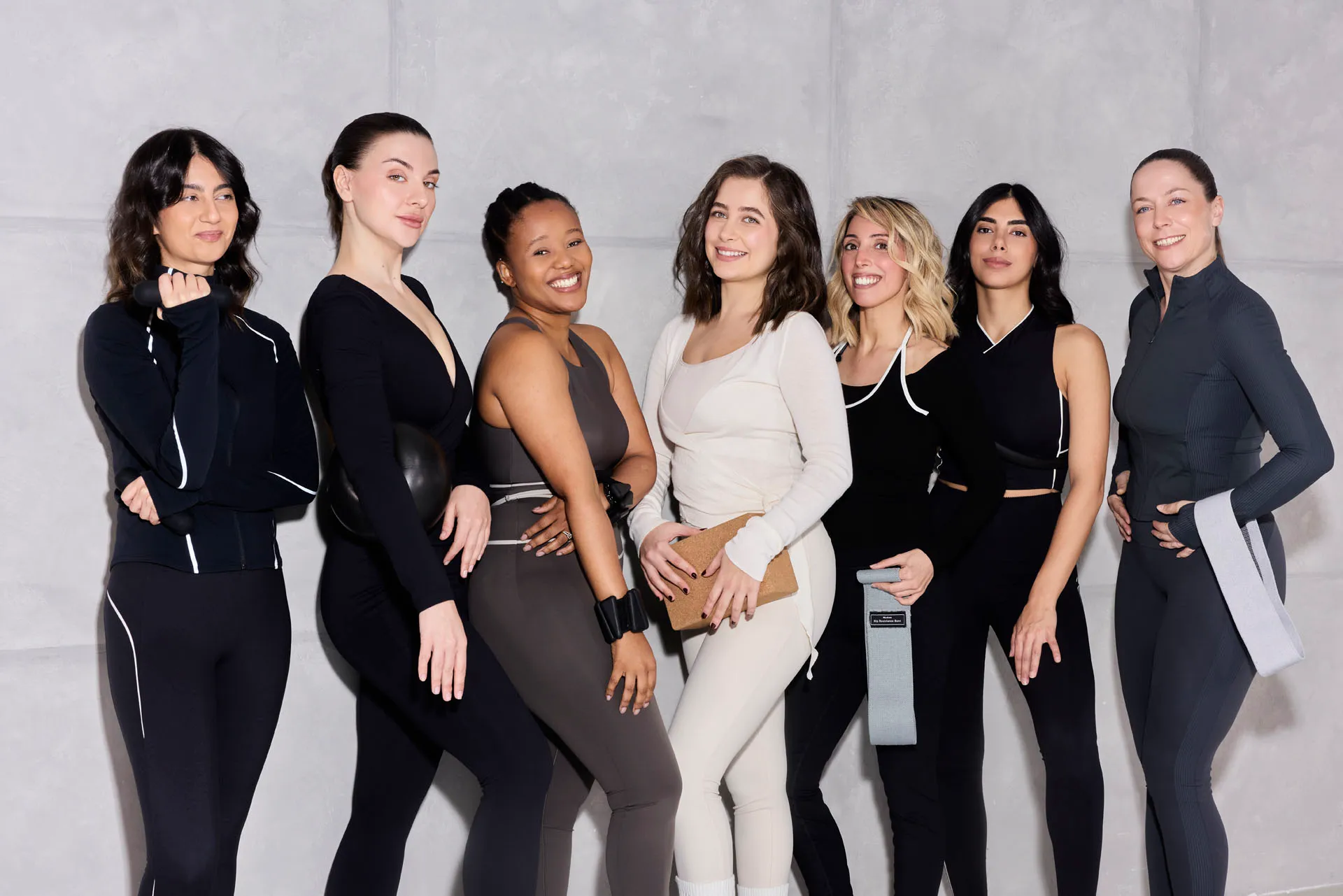 Seven women in stylish activewear pose in front of a gray wall, holding fitness accessories.