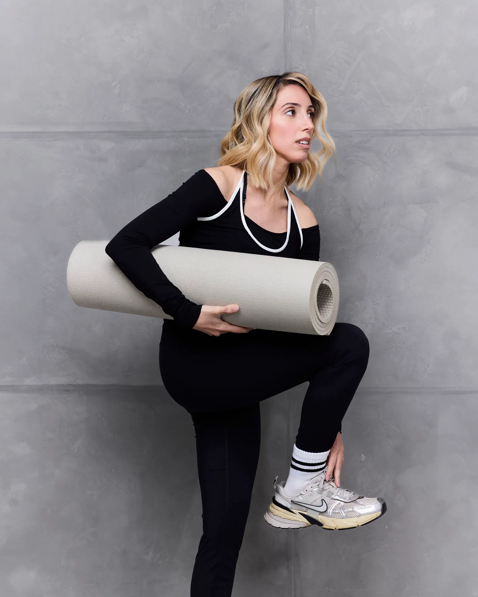 Woman in black athletic wear holding a rolled yoga mat against a gray wall.