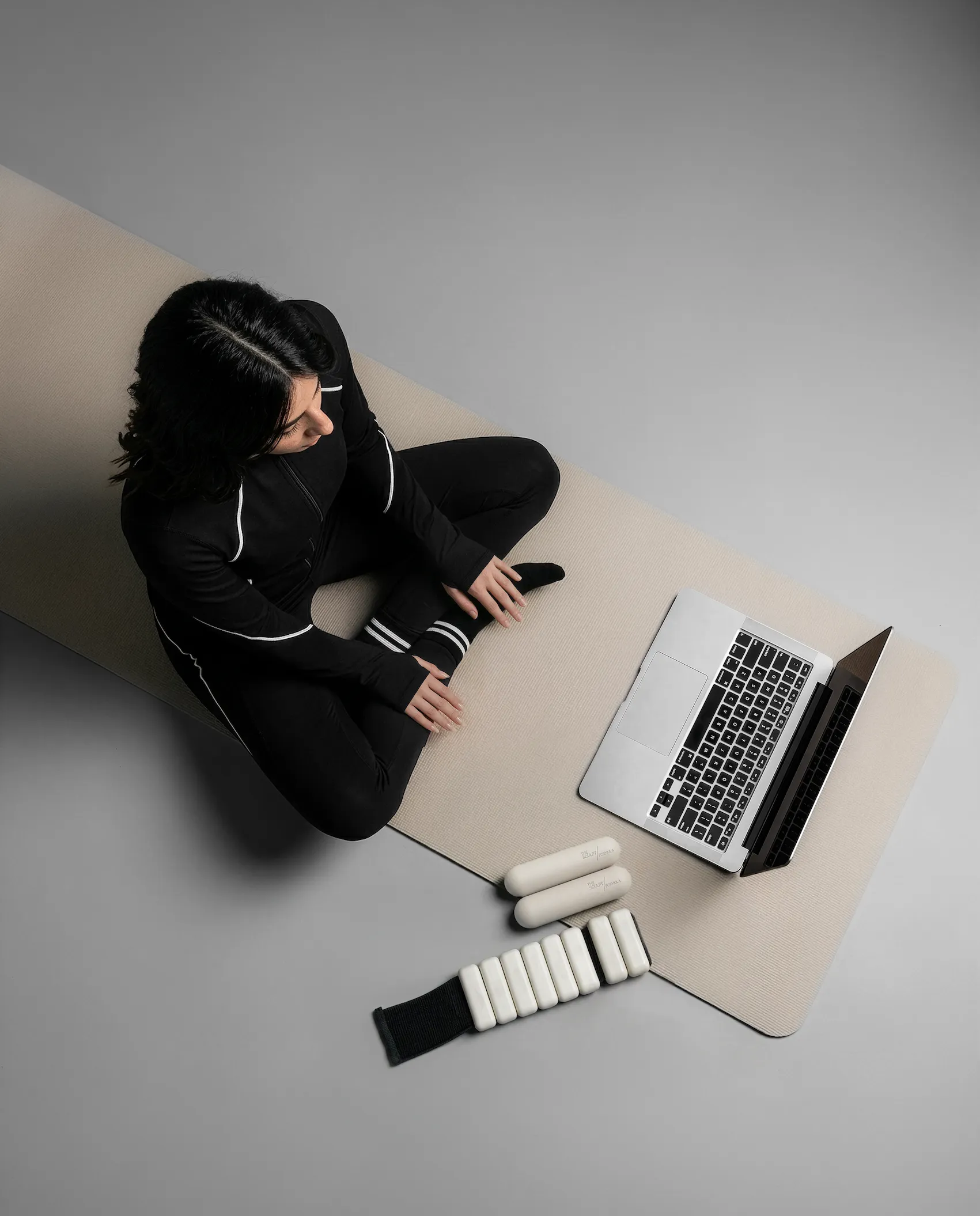 Woman in black workout clothes sitting cross-legged on a beige mat next to a laptop and weights.