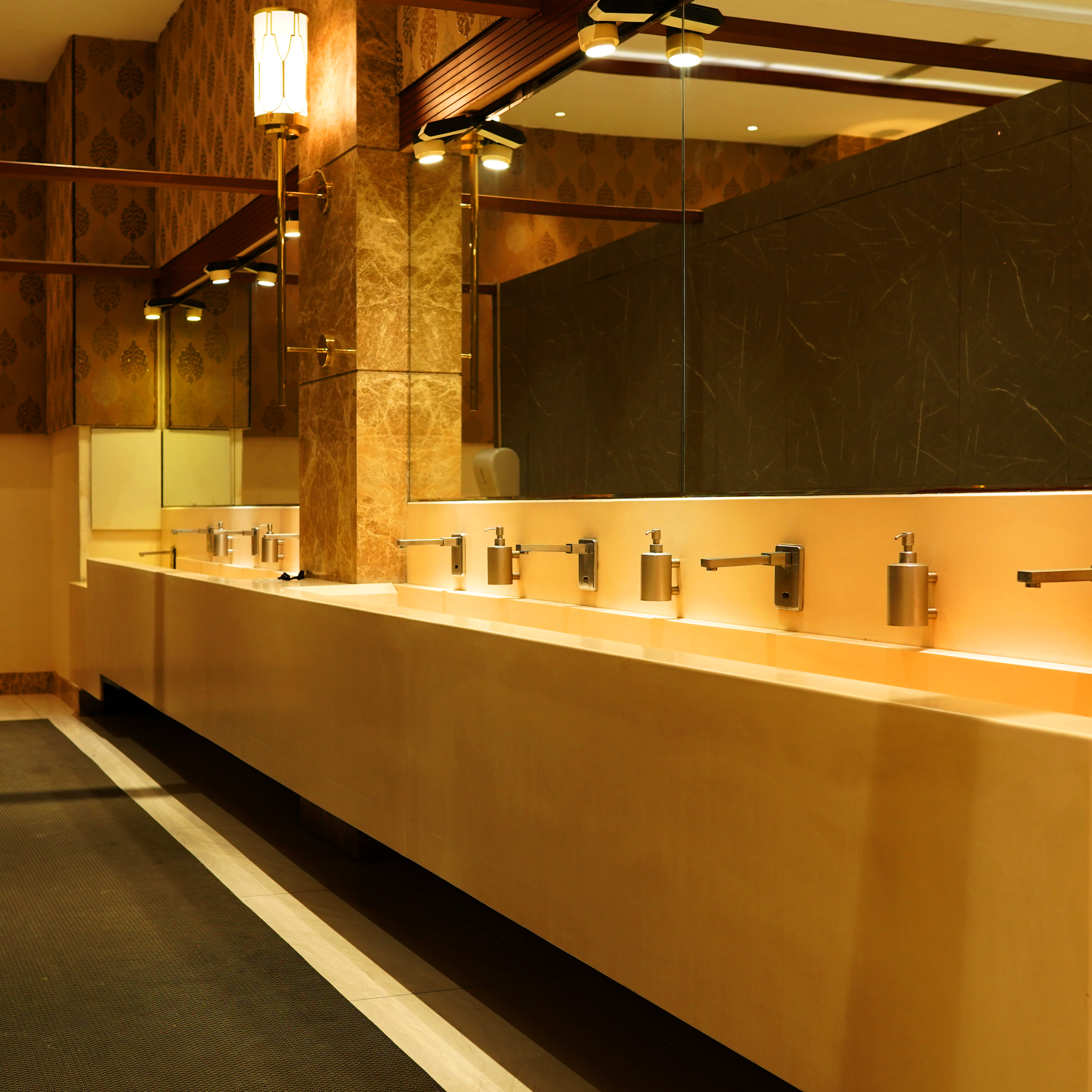 Luxurious restroom with long beige countertop featuring multiple stainless steel faucets and soap dispensers under warm lighting.