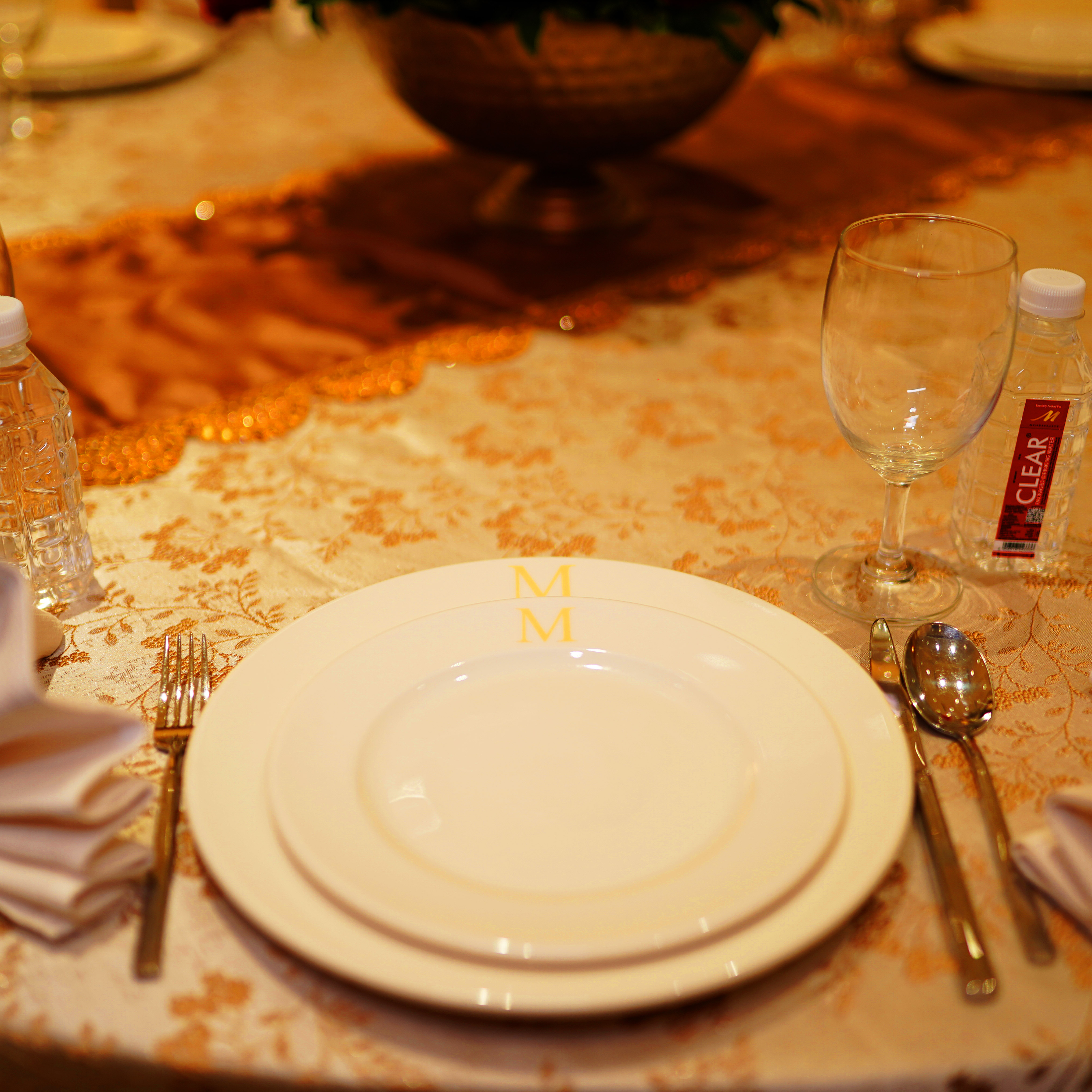 Elegant table setting with two stacked white plates featuring gold initials 'M', fork on left, spoon and knife on right, wine glass, water bottles, and folded napkins on patterned tablecloth.