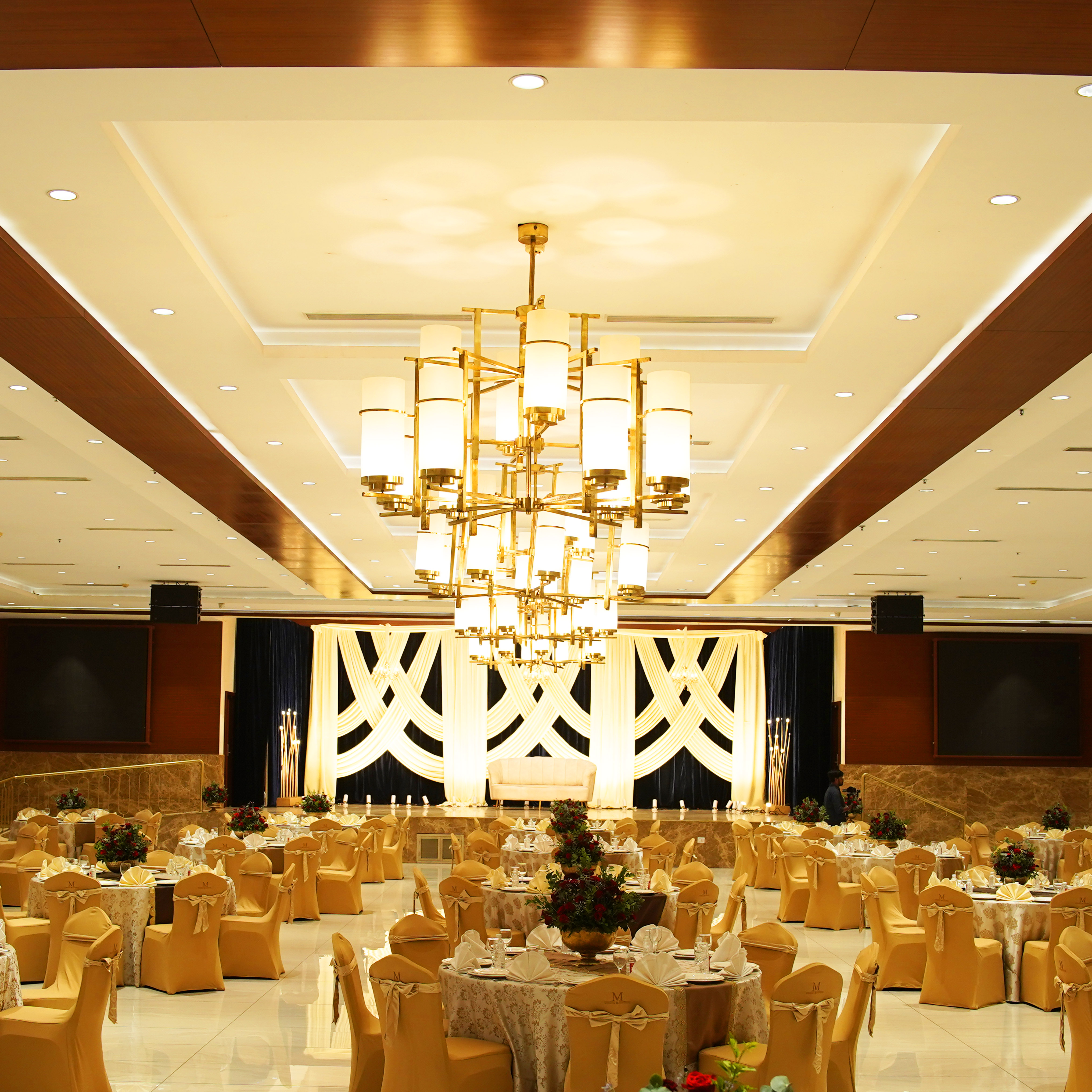Elegant banquet hall with round tables covered in floral tablecloths and gold chairs, a large modern chandelier, and a decorated stage with draped white fabric.