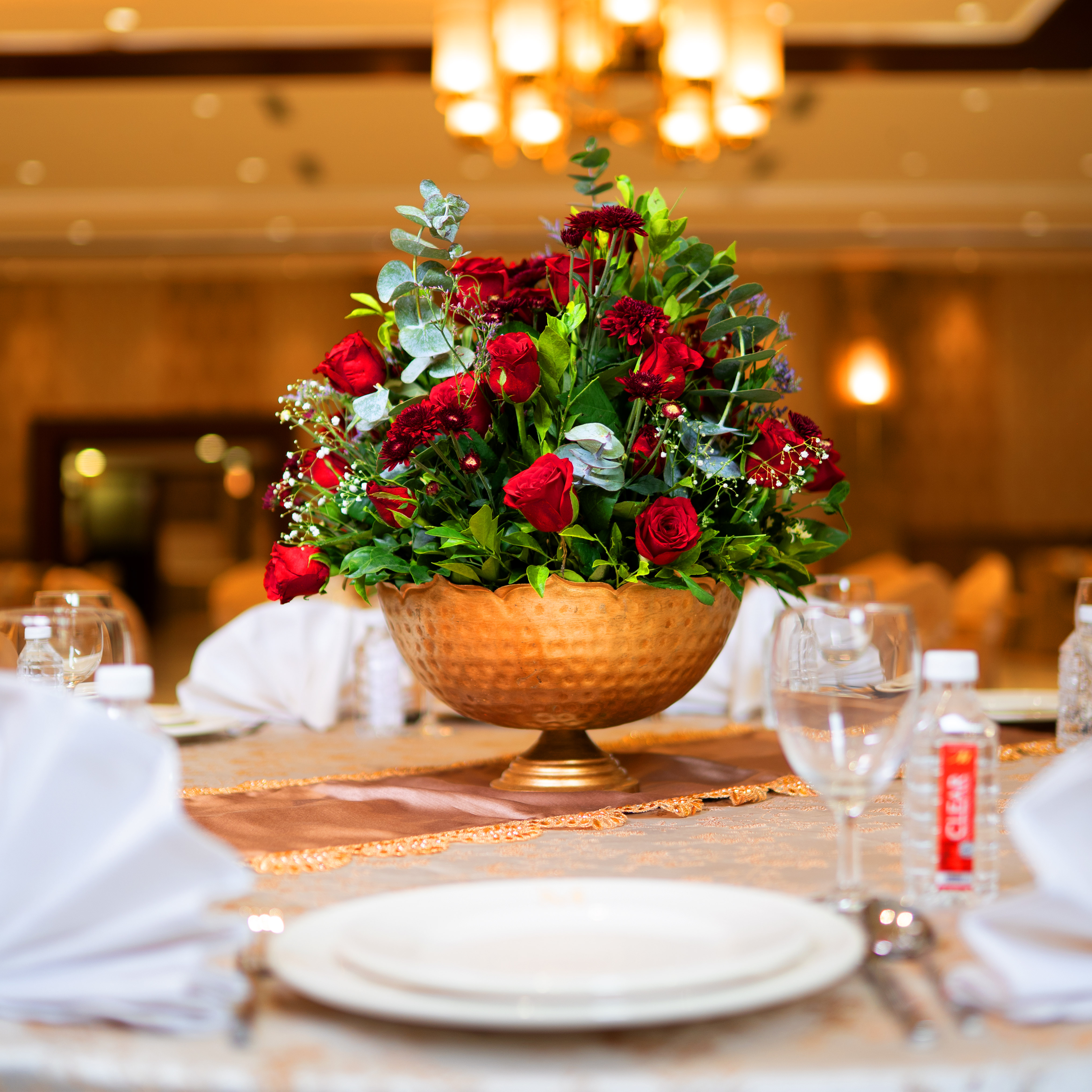 Elegant floral centerpiece with red roses and greenery in a hammered gold vase on a set dining table.
