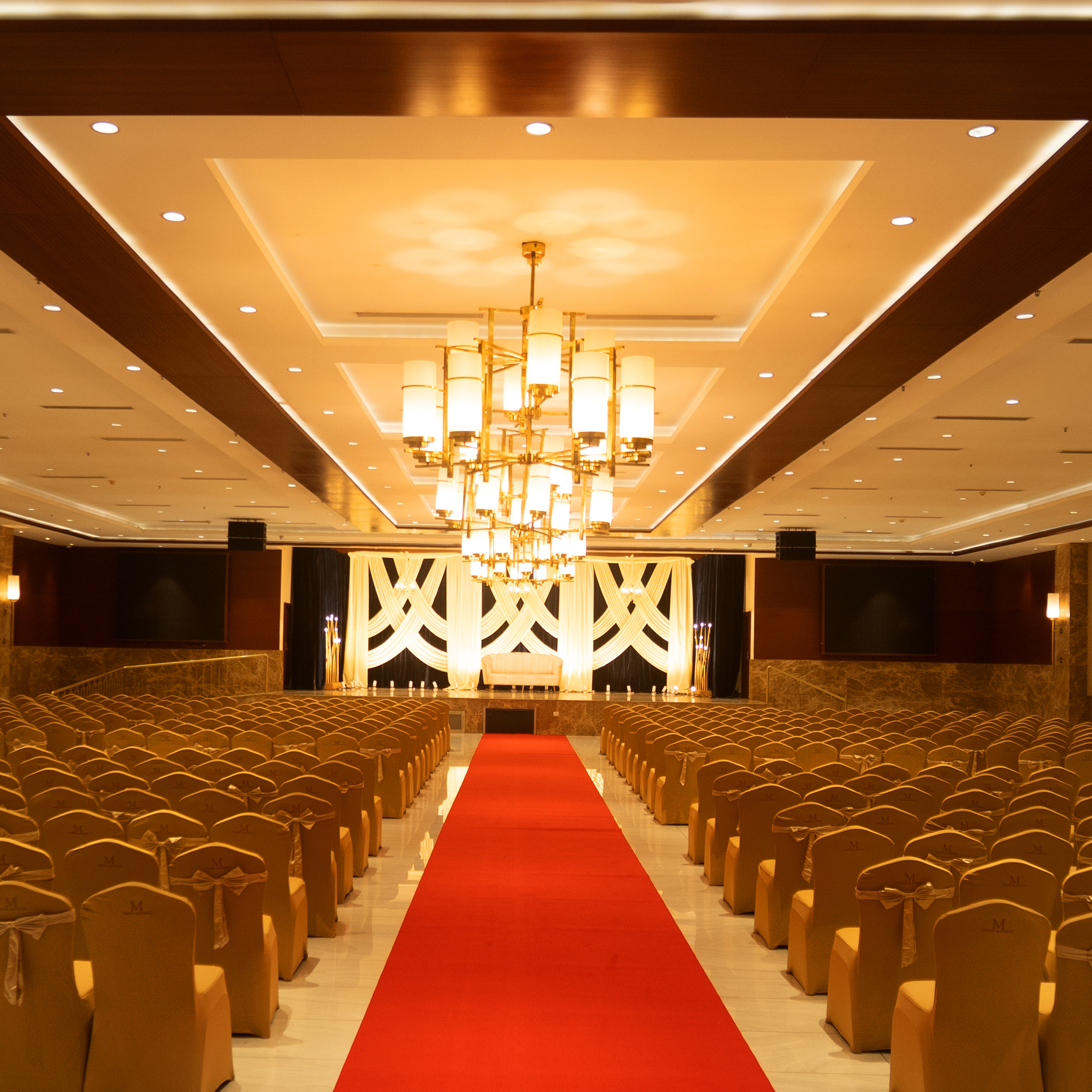 A spacious event hall with rows of gold-covered chairs on either side of a red carpet leading to a stage with white draped curtains and a sofa under large chandeliers.