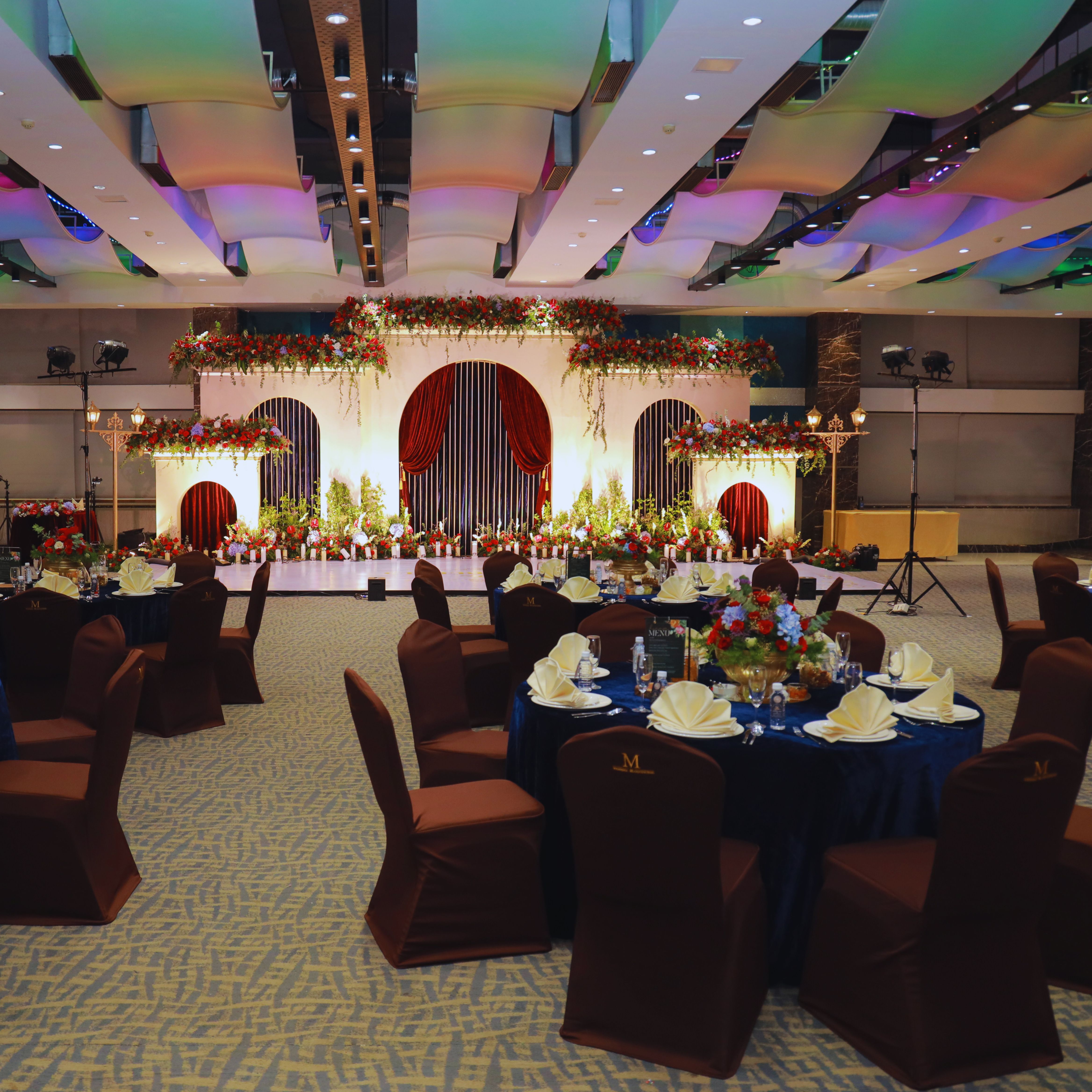 Banquet hall with round tables set with white napkins, floral centerpieces, and a decorated stage with red curtains and flowers.