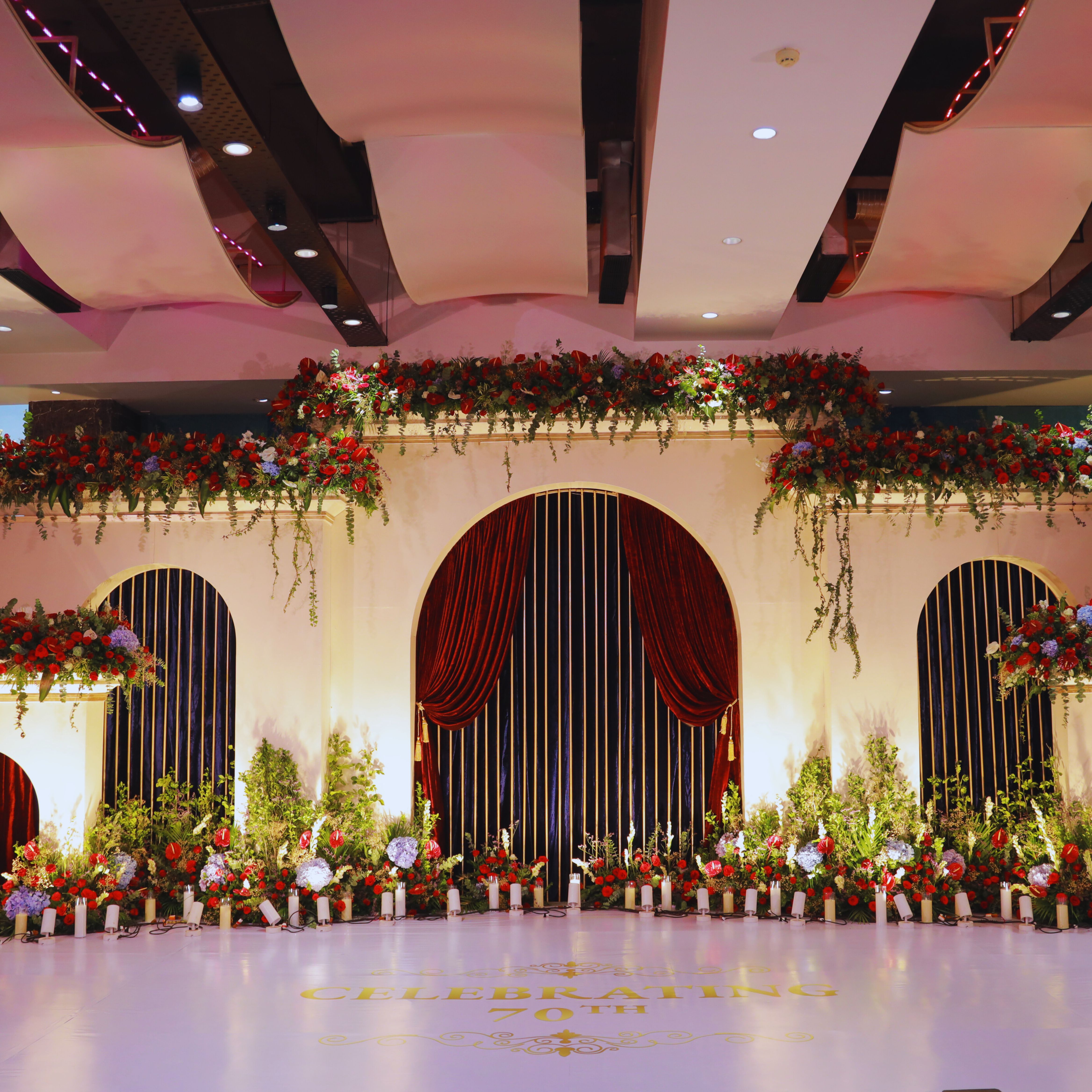 Elegant event stage with large cream arches, red velvet curtains, lush floral arrangements of red and purple flowers, and candles, celebrating a 70th milestone.