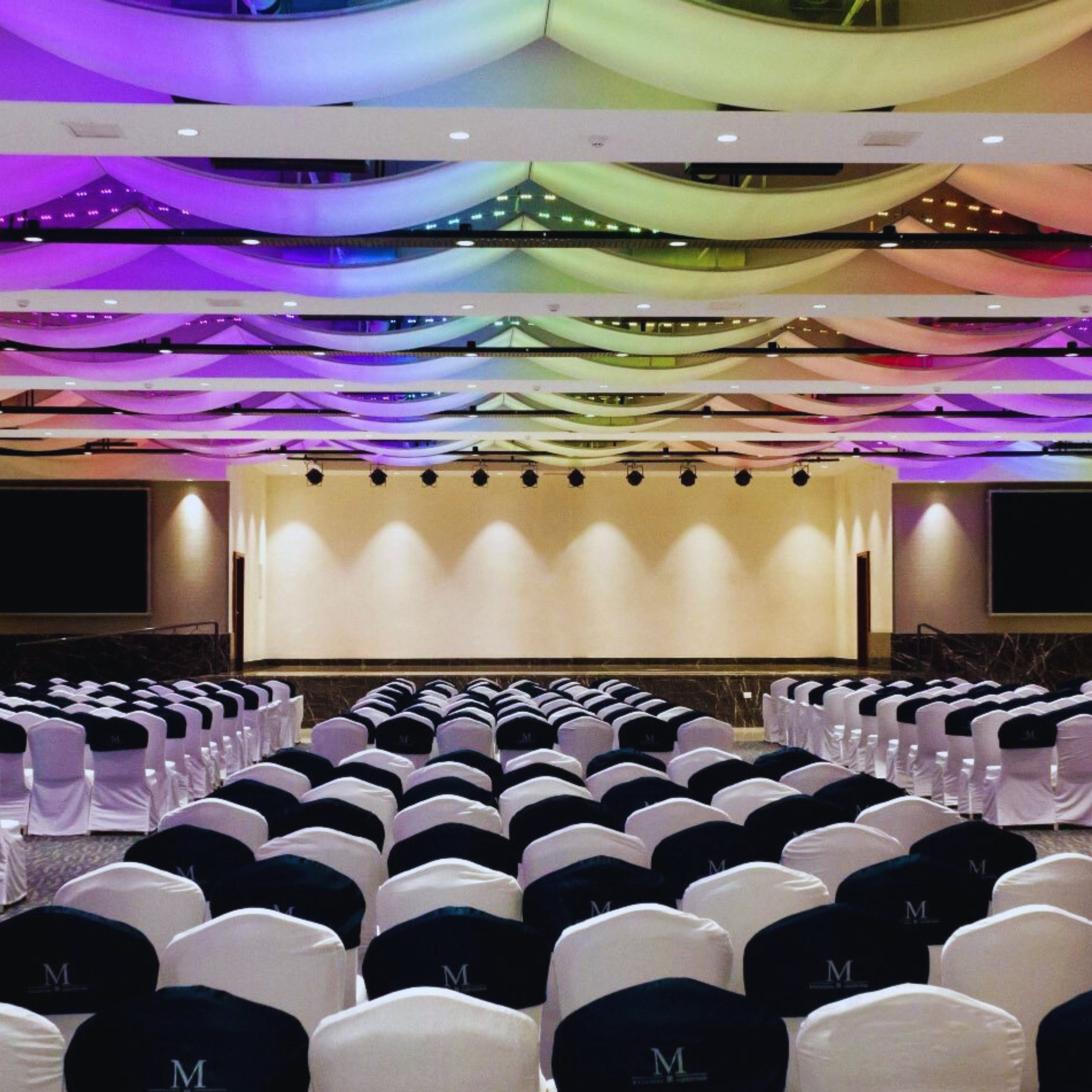 Conference room with rows of white chairs covered in black and white seat covers facing an empty stage, ceiling decorated with colorful draped fabric and lights.