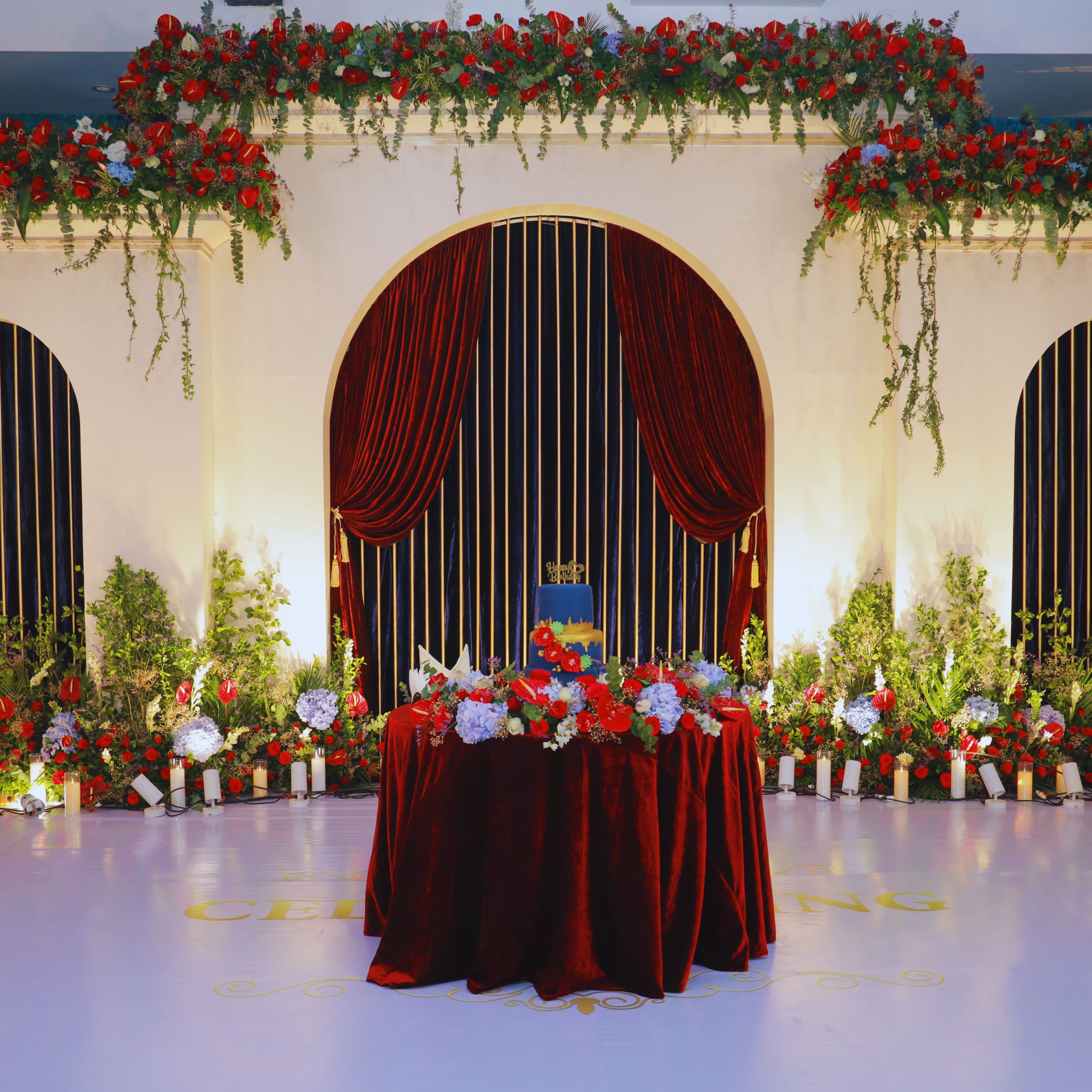 Elegant birthday party setup with a blue and gold cake on a red velvet tablecloth surrounded by red and blue floral arrangements and candles.
