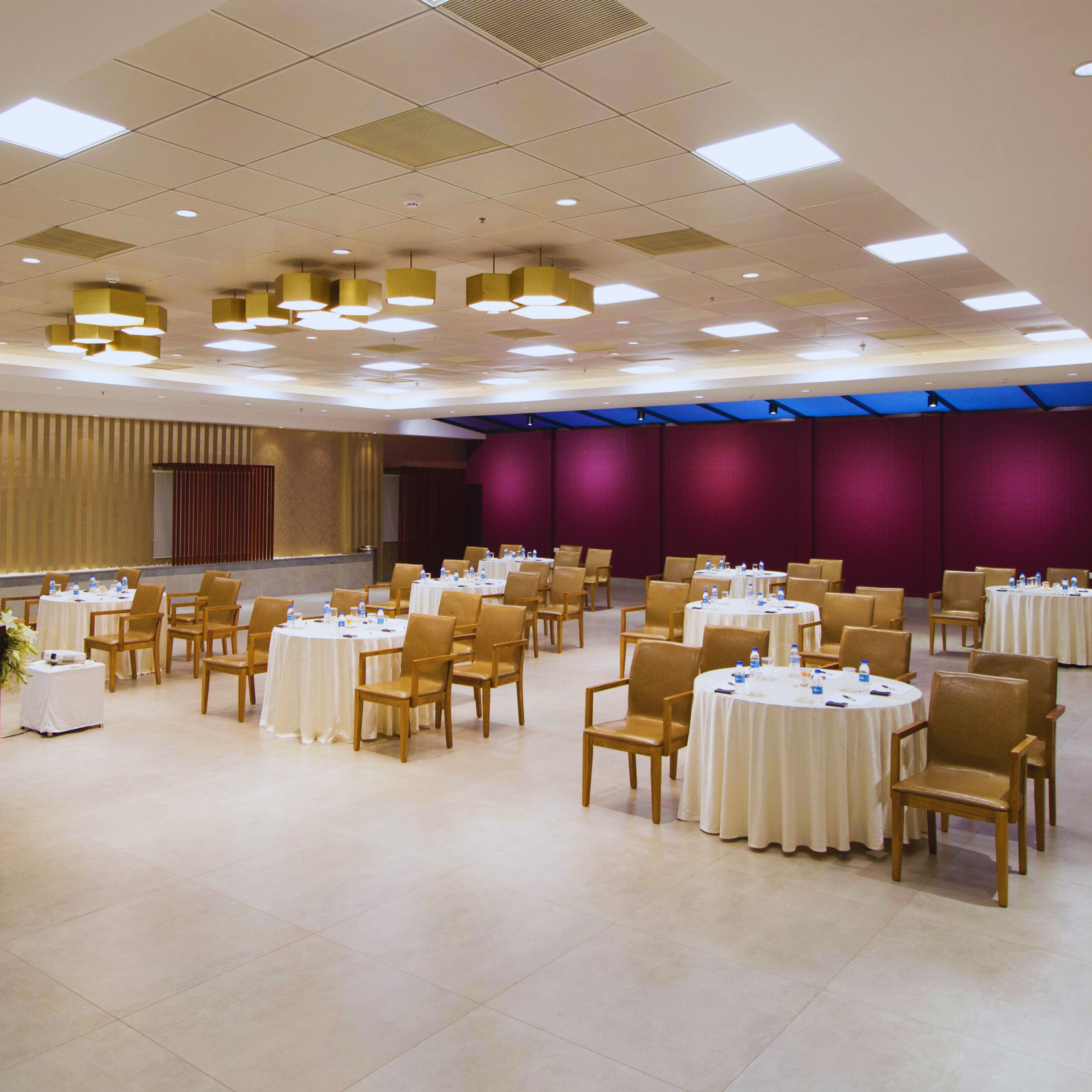 Spacious banquet hall with round tables covered in white tablecloths, brown chairs, and ceiling lights.