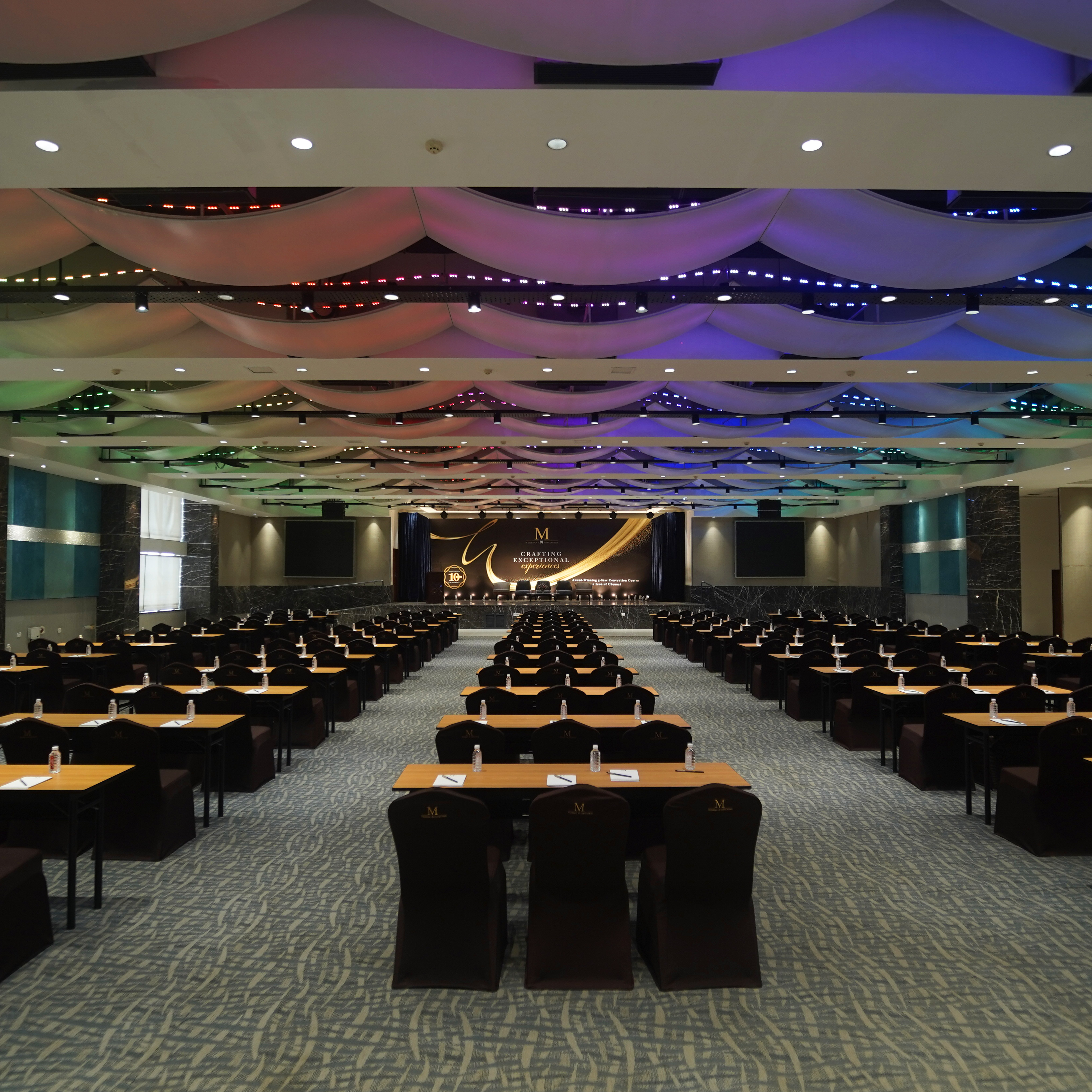 Large conference room with rows of tables and chairs facing a stage with a decorative ceiling featuring multicolored lighting.
