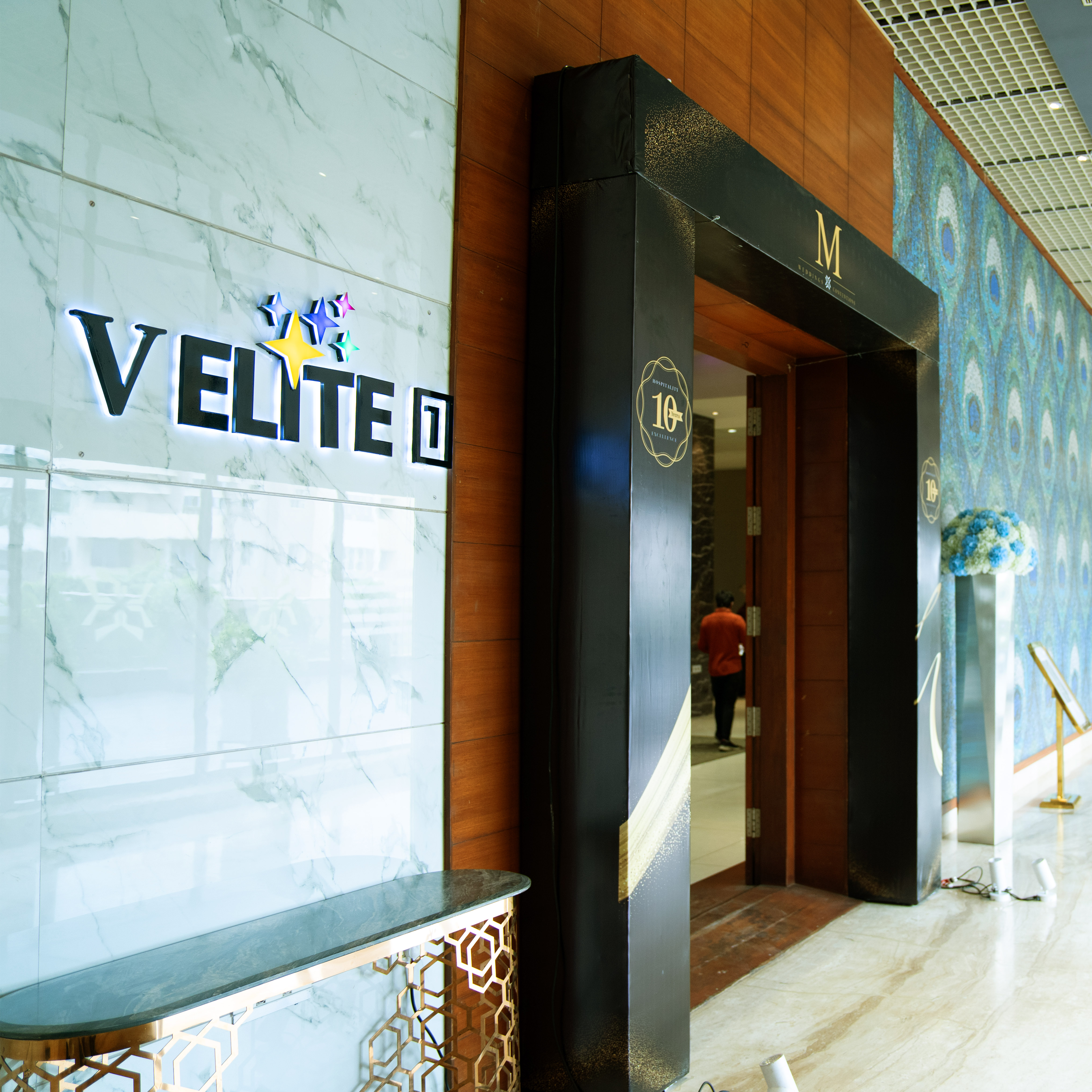 Entrance to a venue with a lit sign reading 'VELITE' on a marble wall and a black archway marked with '10' and a flower arrangement on the right.