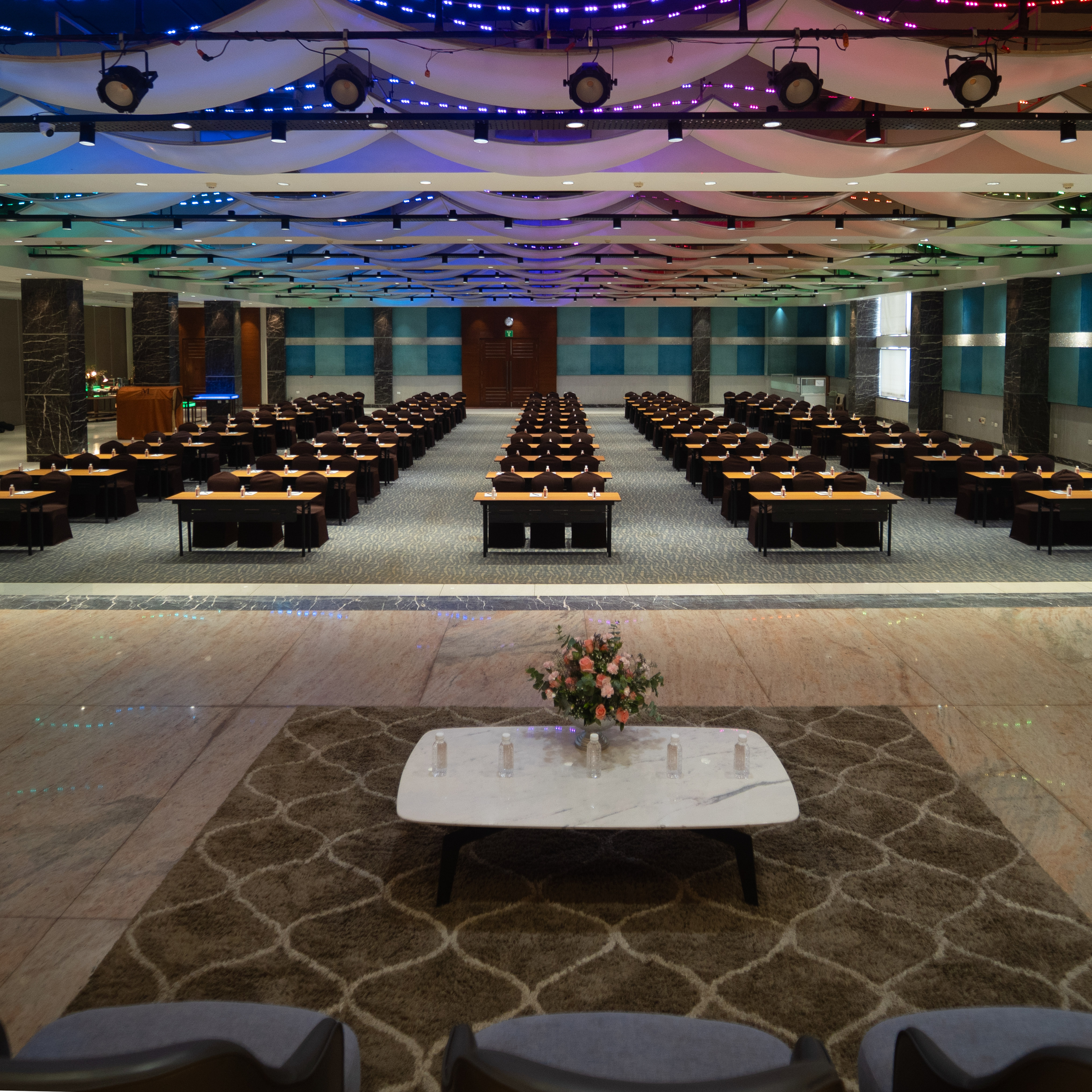 Large conference room arranged with rows of tables and chairs facing a stage with a coffee table and flower arrangement.