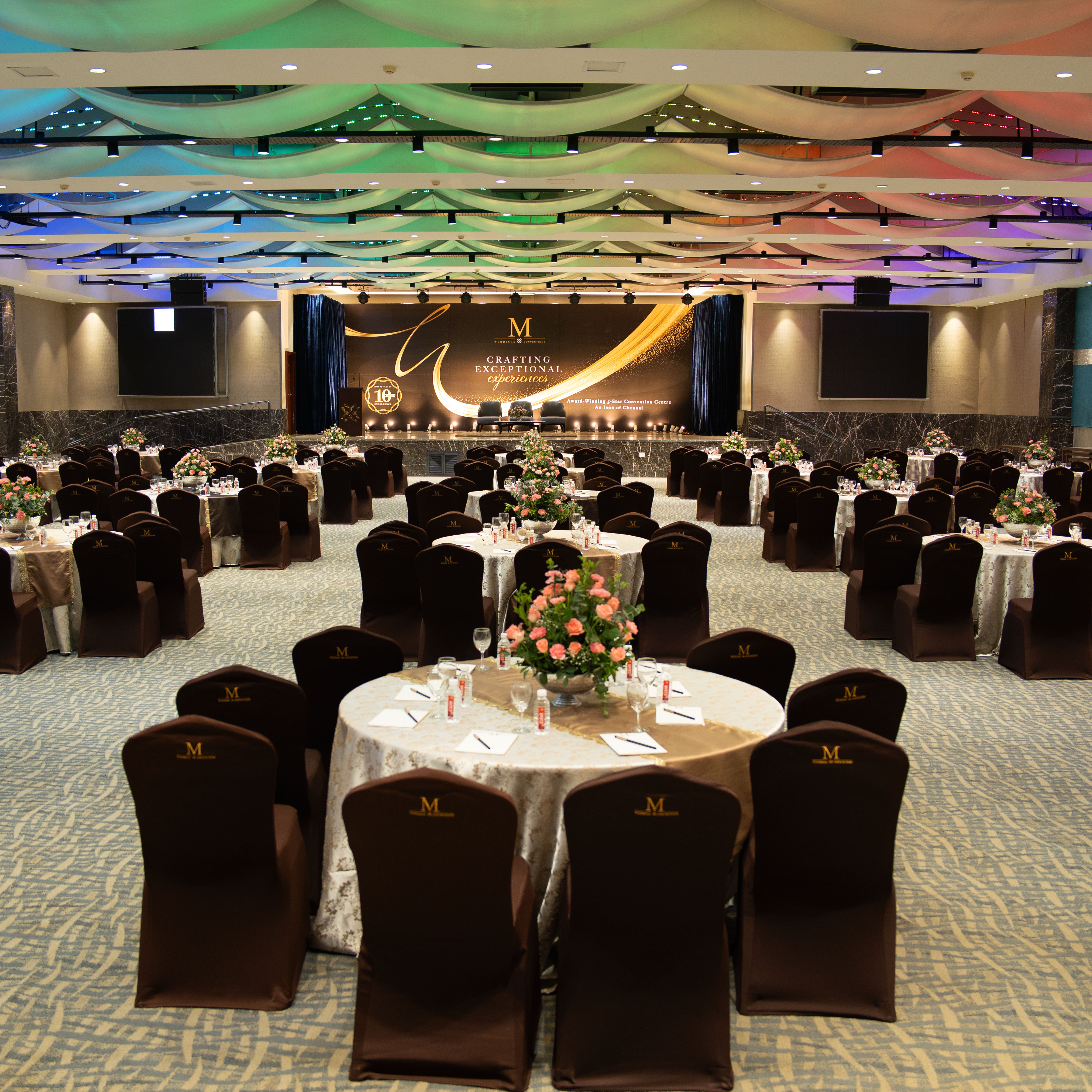 Elegant banquet hall with round tables set for an event, floral centerpieces, and a stage with a backdrop that reads 'Crafting Exceptional Experiences'.