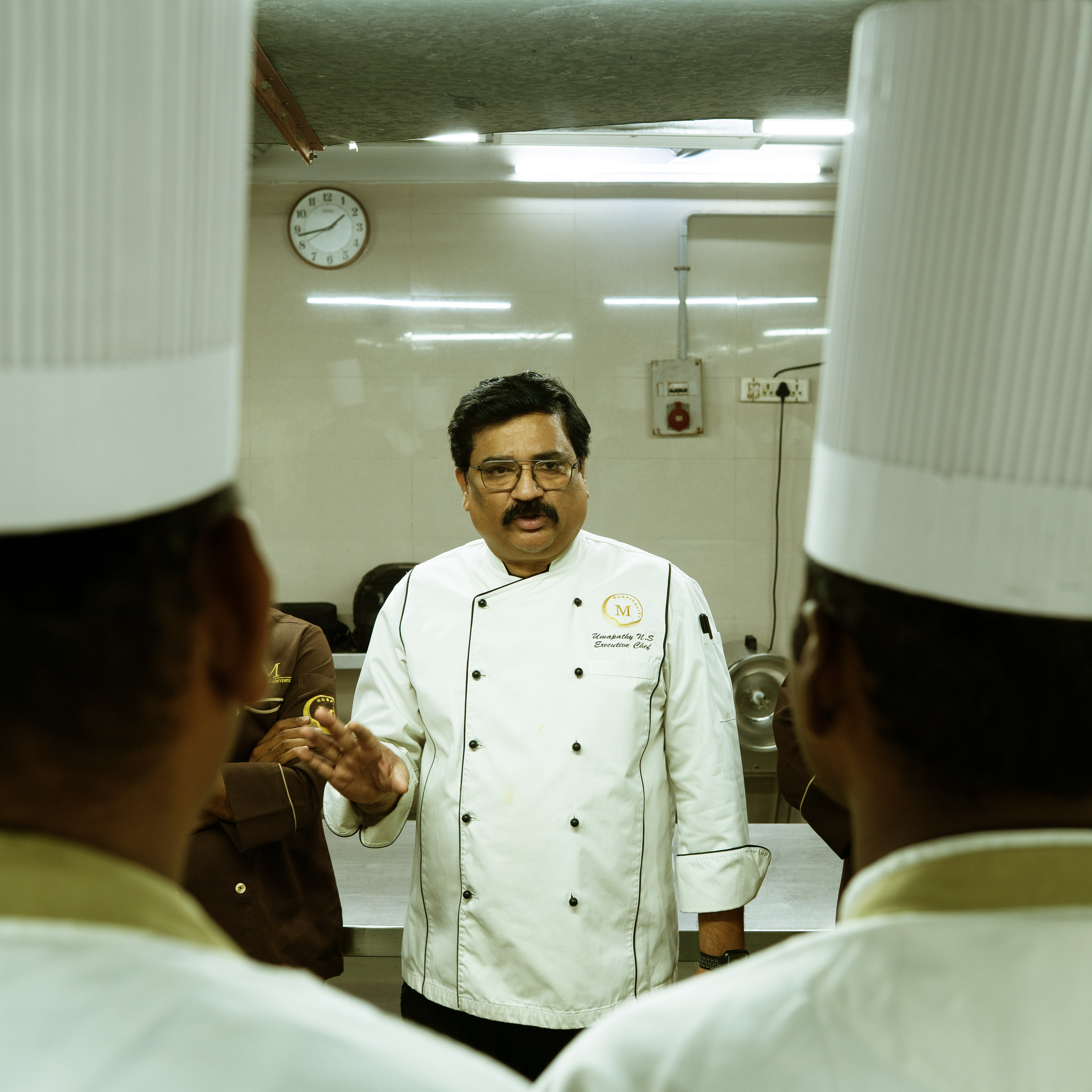 Executive chef speaking to two chefs wearing tall white hats in a professional kitchen.