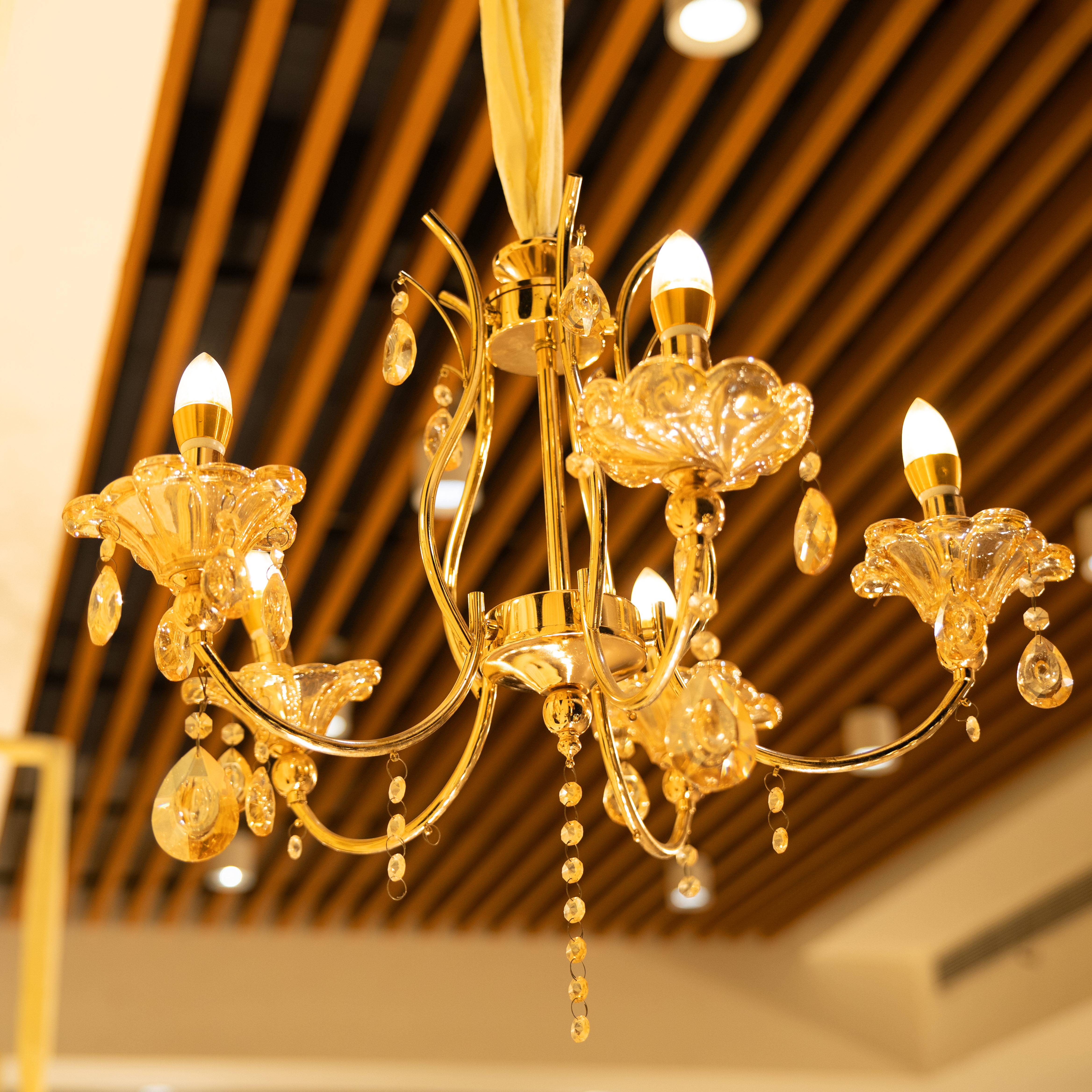 Ornate gold chandelier with crystal decorations and five glowing lights hanging from a wooden slatted ceiling.