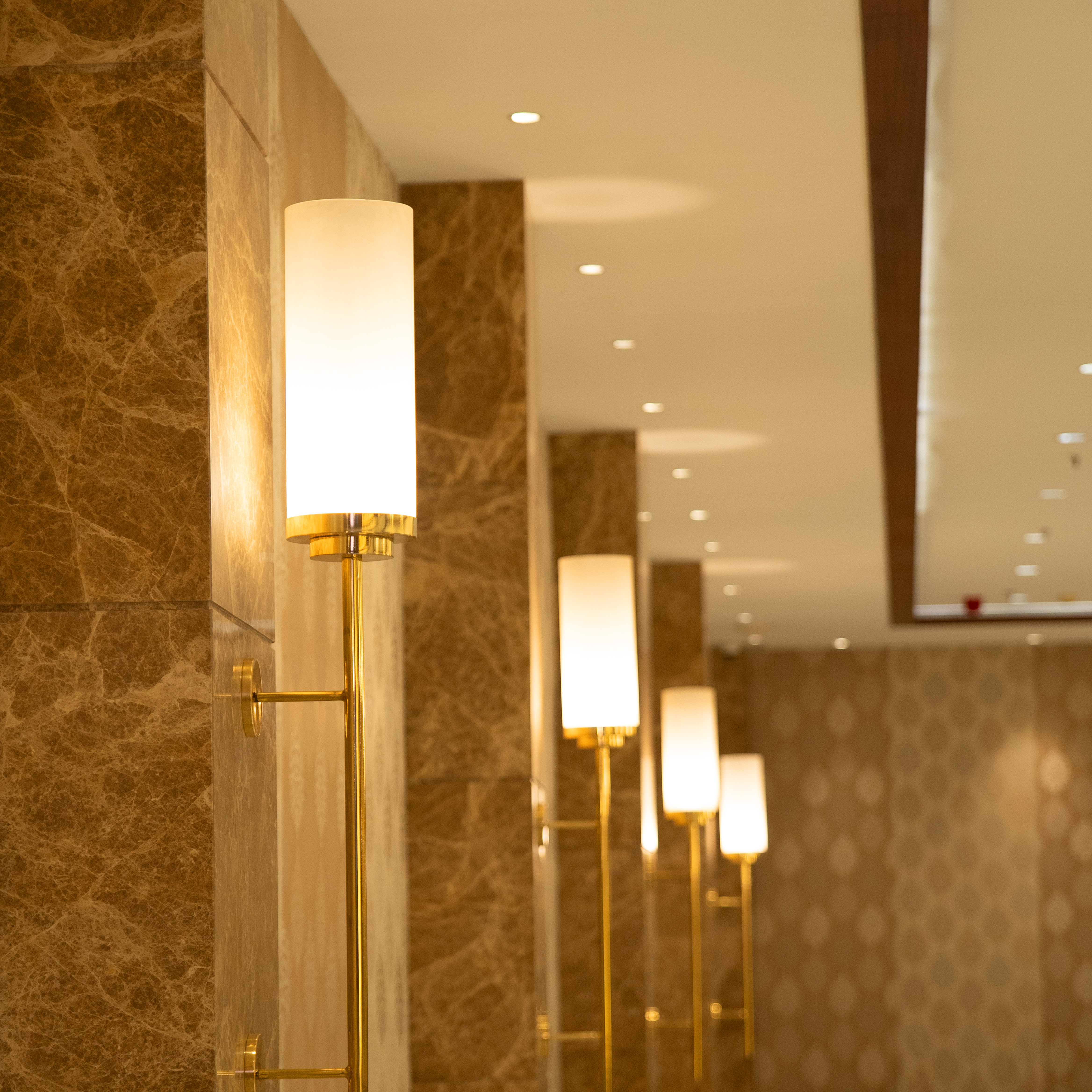 Row of modern wall lamps with cylindrical frosted glass shades and brass fixtures on marble walls in a hallway.