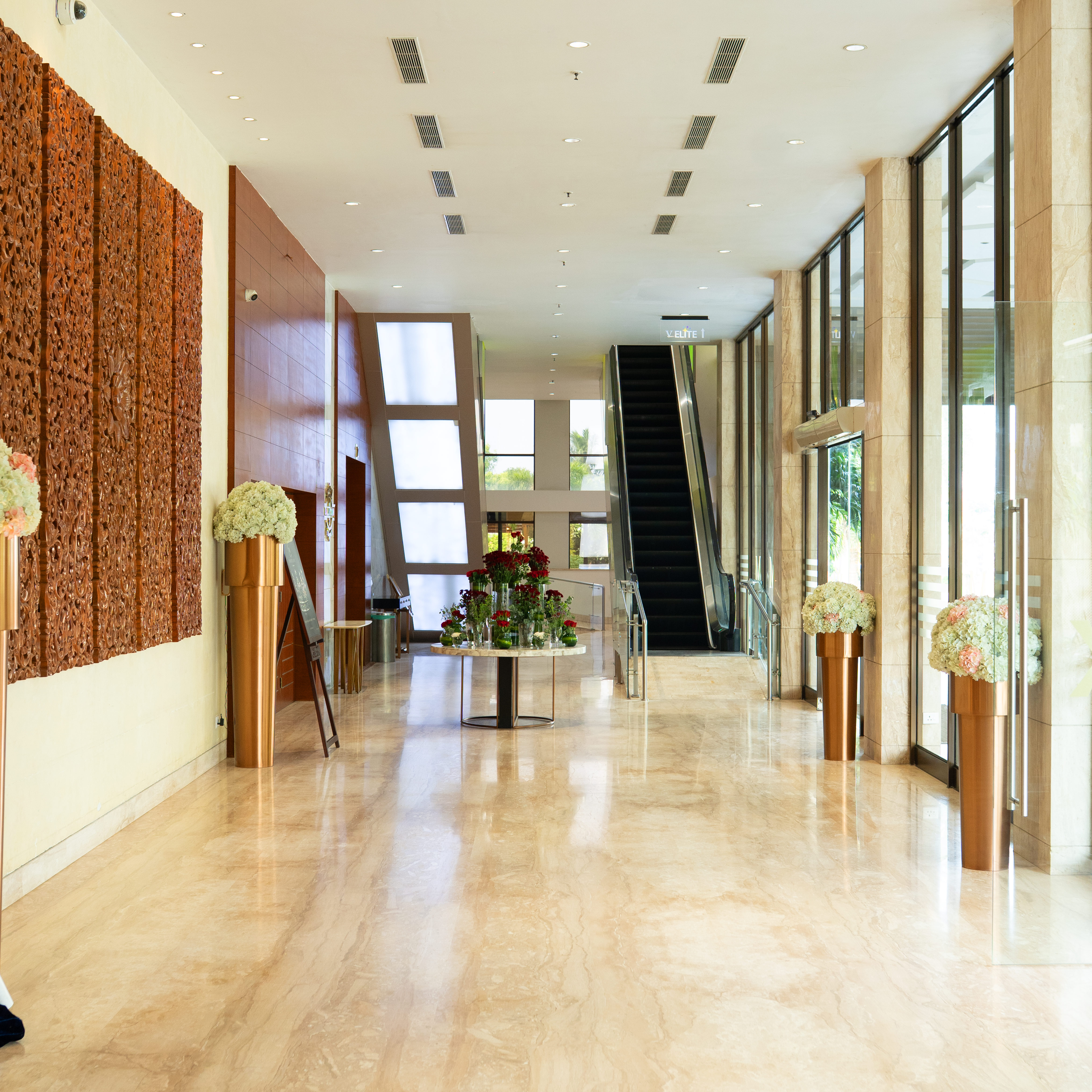 Bright hotel lobby with marble floors, tall flower arrangements, carved wooden wall art, and an escalator in the background.