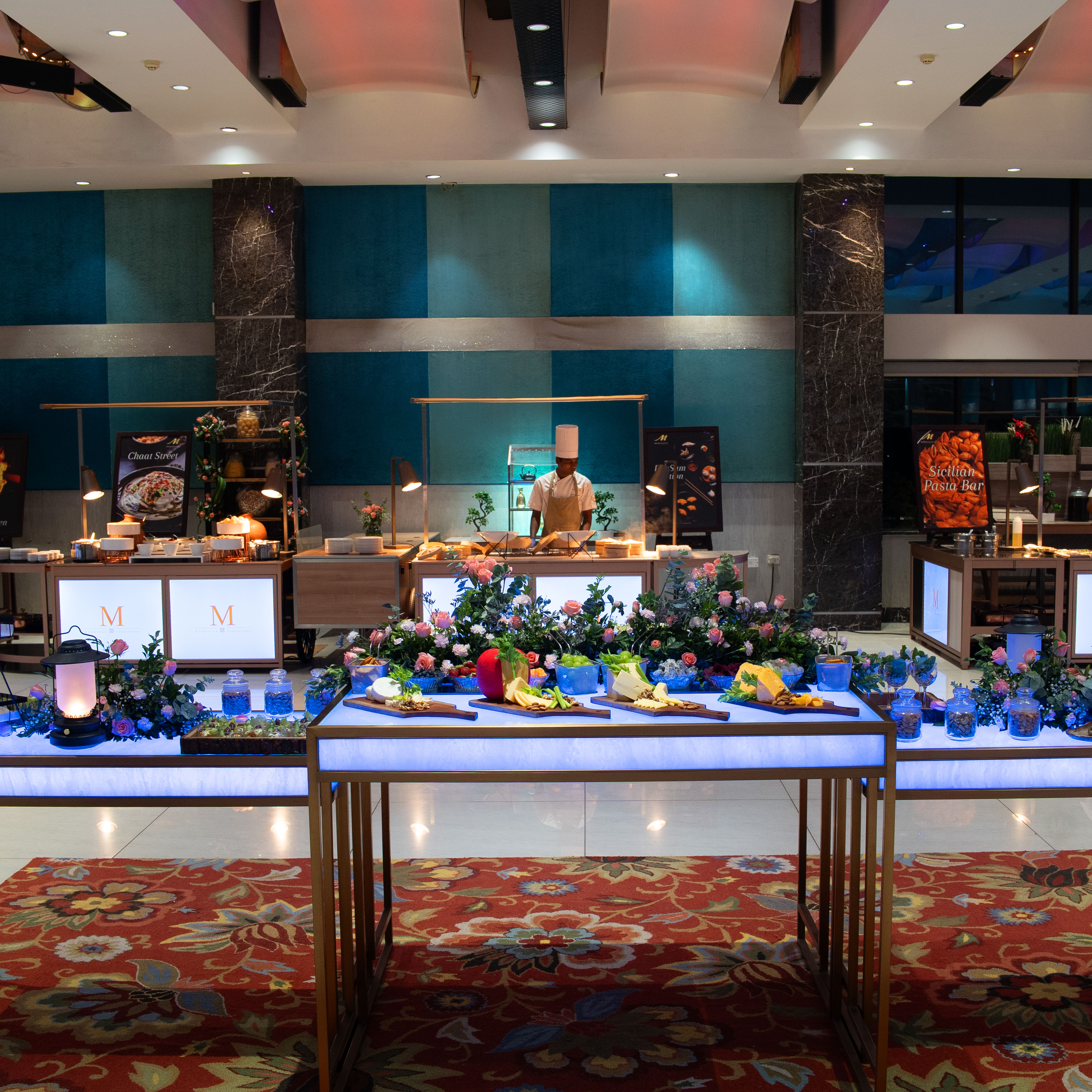 Chef standing behind a buffet table decorated with flowers and various food dishes in a well-lit indoor setting.
