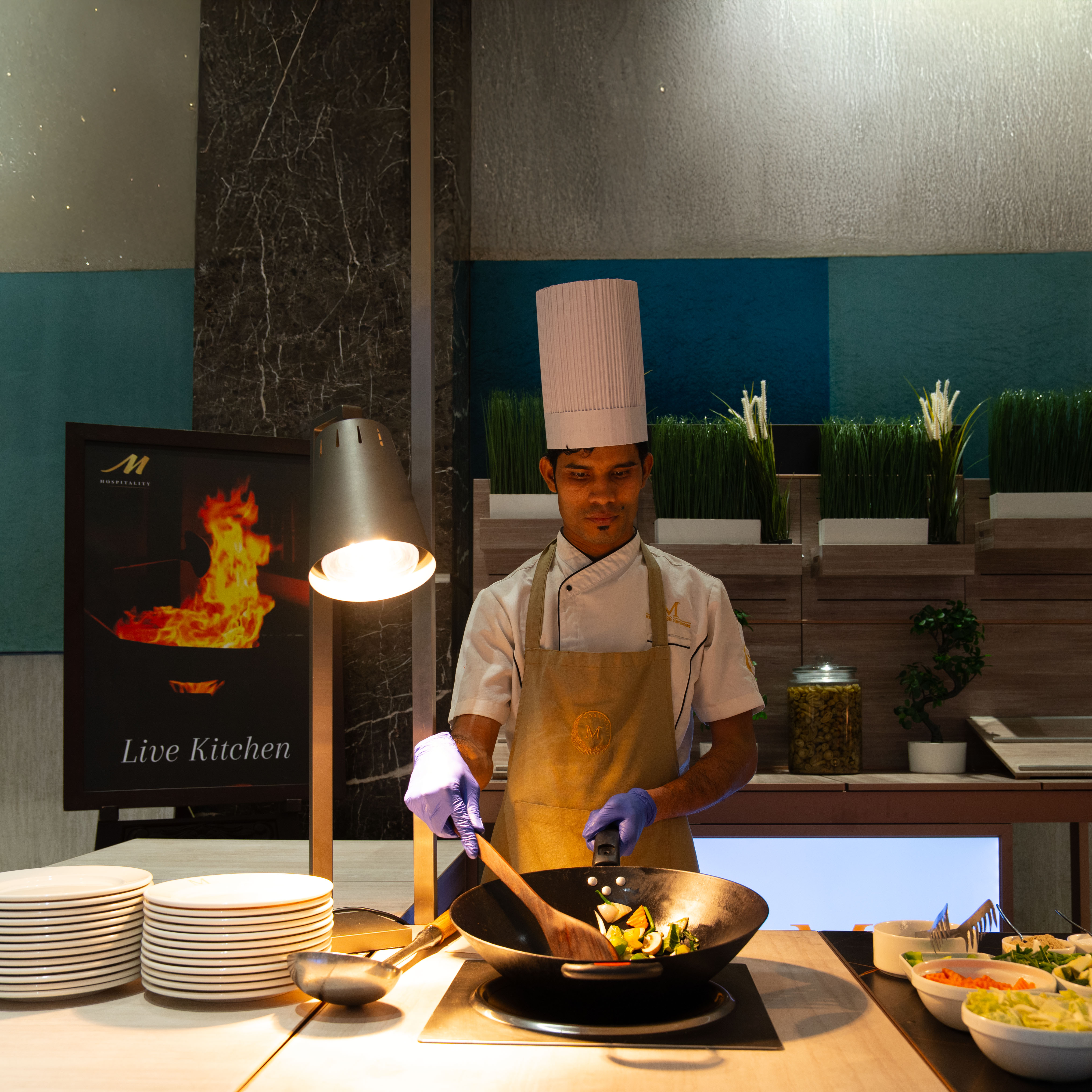 Chef in white uniform and tall hat cooking vegetables in a wok at a live kitchen station with plates and salad bowls nearby.