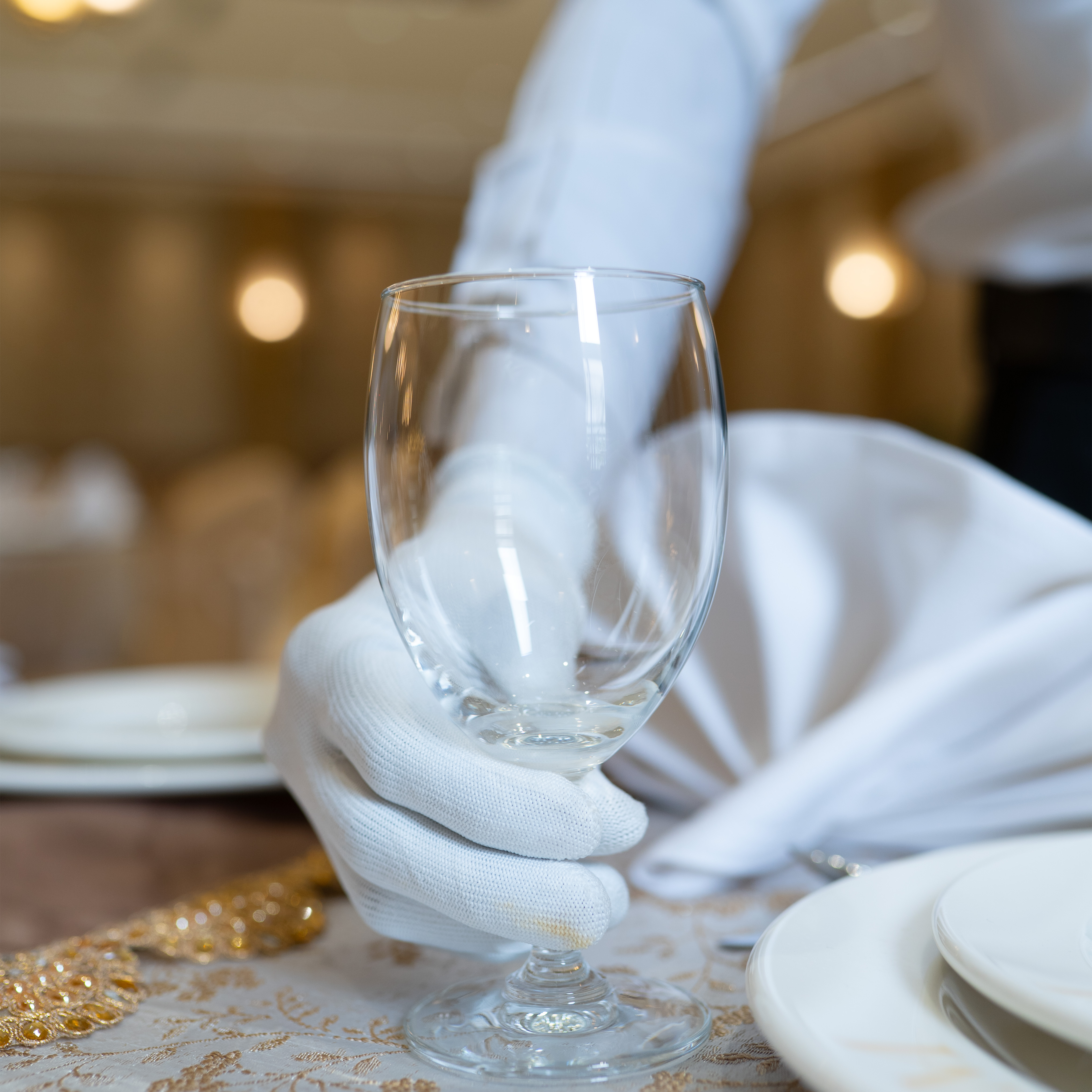 Person wearing white gloves placing a clear empty wine glass on a decorated table with white plates and napkins.