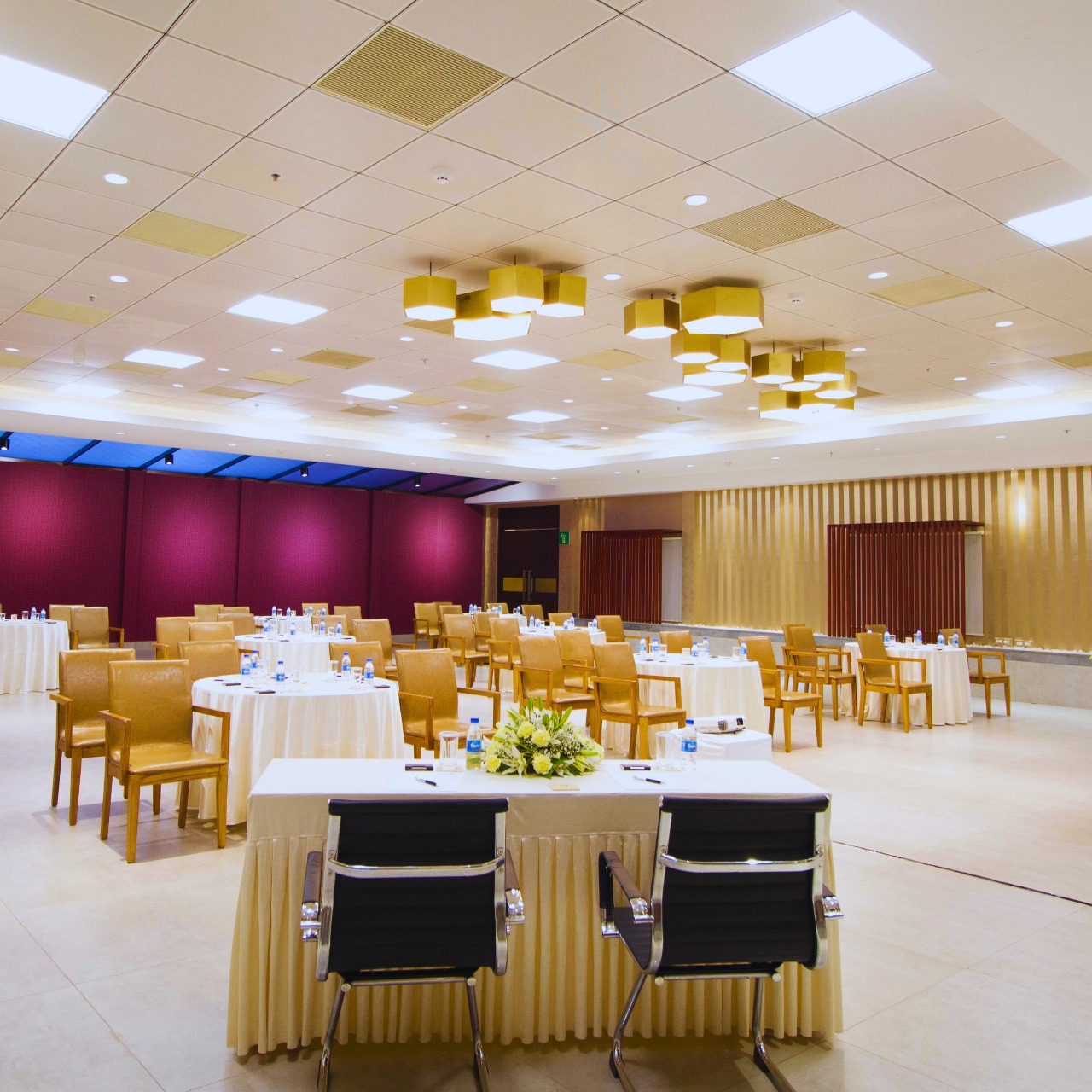 Spacious conference room with round tables covered in white cloth, brown chairs, and a head table with two black chairs and a flower arrangement.
