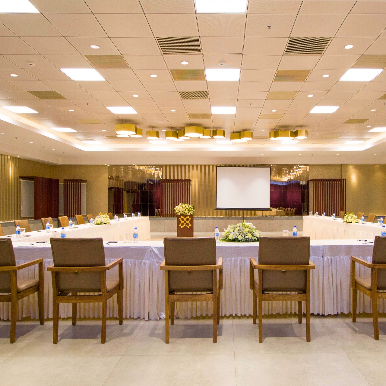 Modern conference room with U-shaped tables, wooden chairs, water bottles, floral centerpieces, and a projection screen.