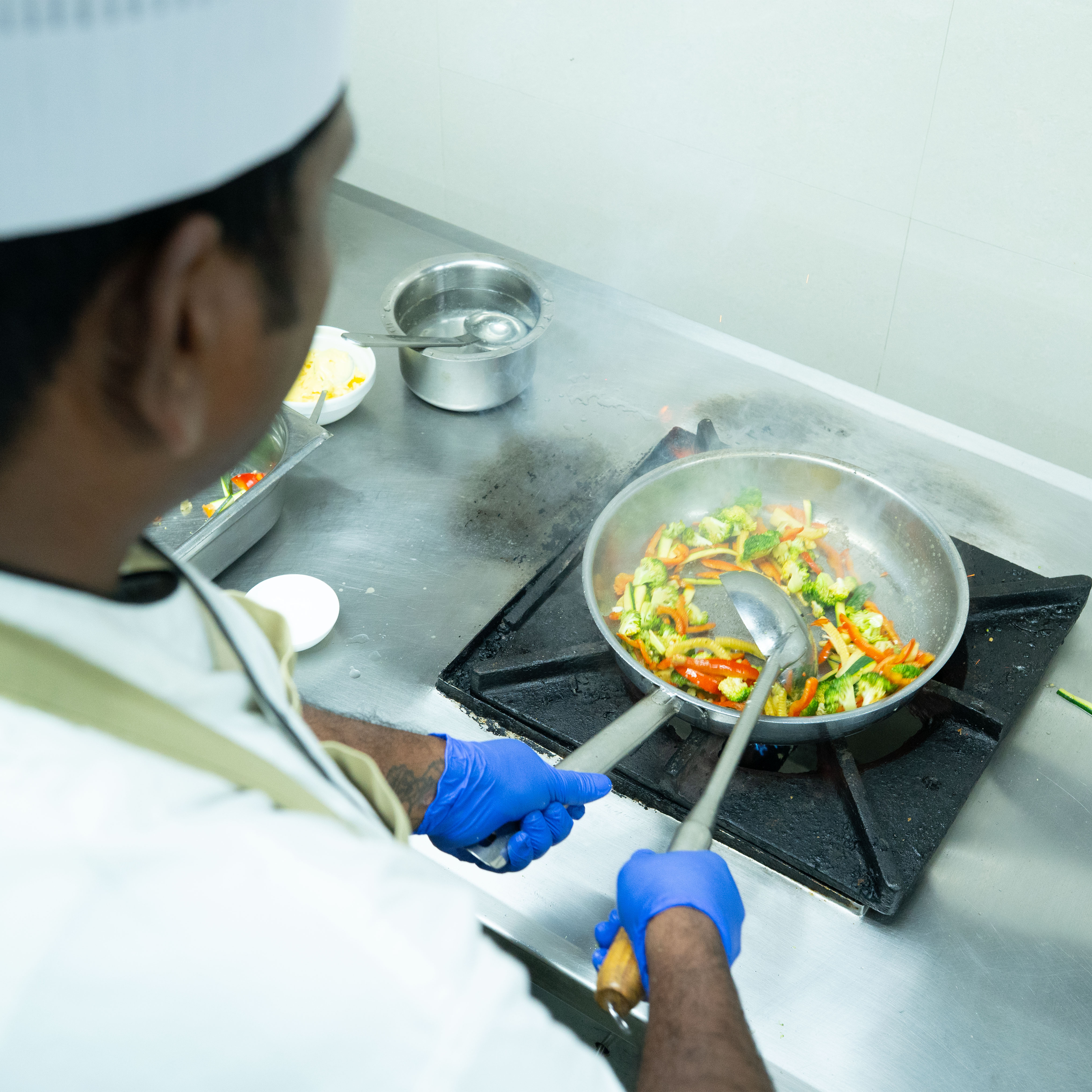 Chef wearing blue gloves stir-frying mixed vegetables including broccoli and bell peppers in a stainless steel pan on a gas stove.